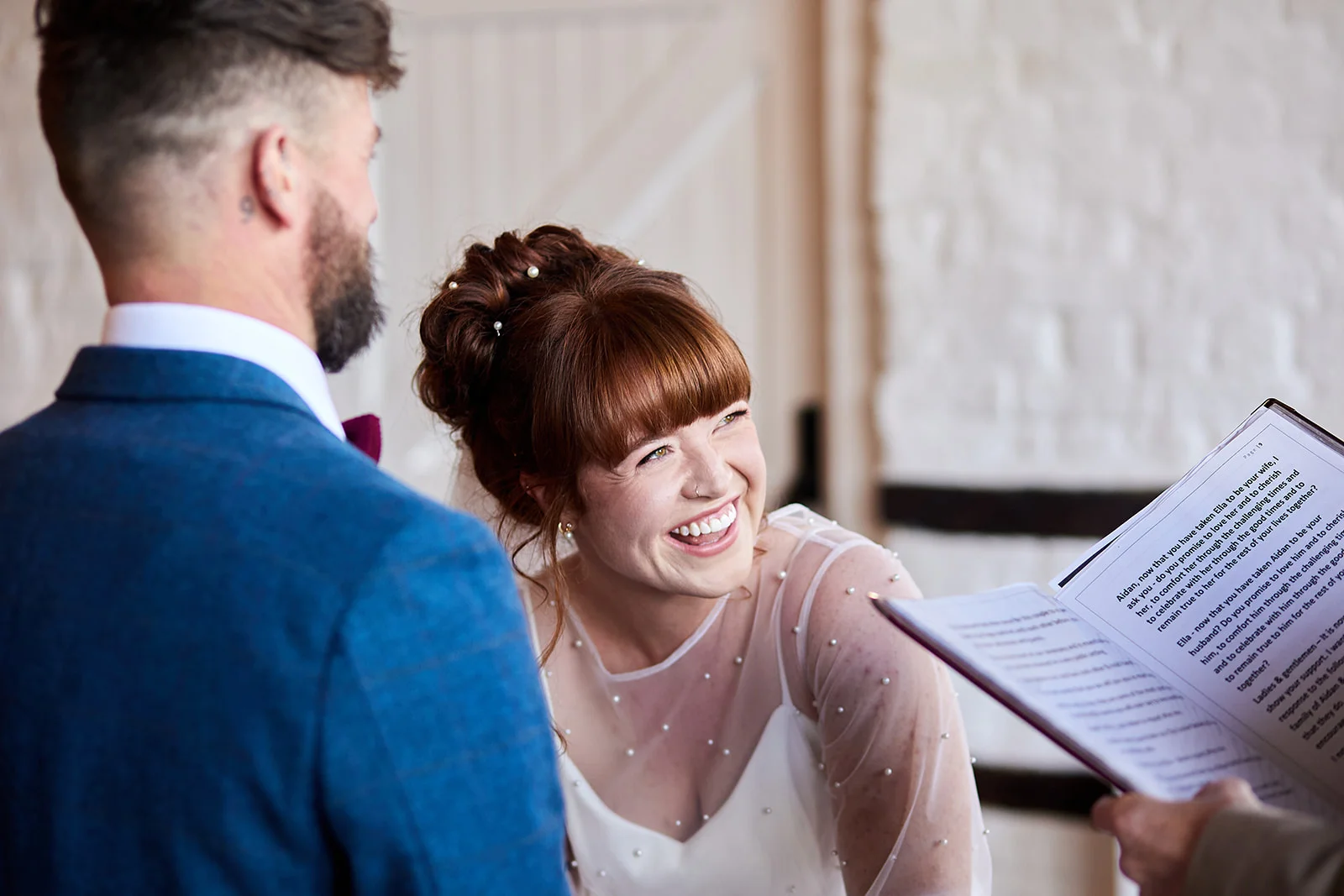 A smiling red-haired bride laughs joyfully during the reading of vows while standing beside her groom in a blue suit inside a rustic barn venue.