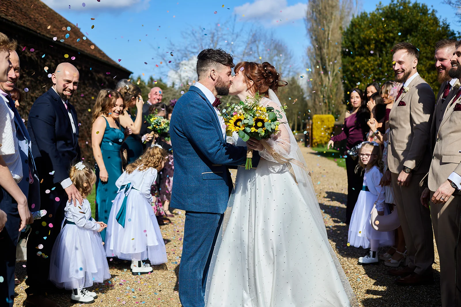 Newlyweds share their first kiss amidst a shower of colourful confetti while guests cheer along a sunlit garden pathway.