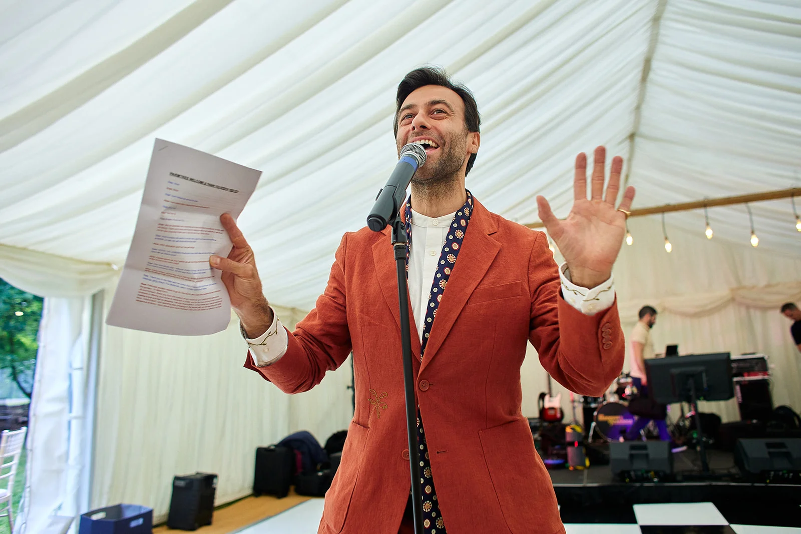 A joyful groom delivers a lively speech inside a marquee, holding speech notes in one hand and raising his other hand while laughing into a microphone.