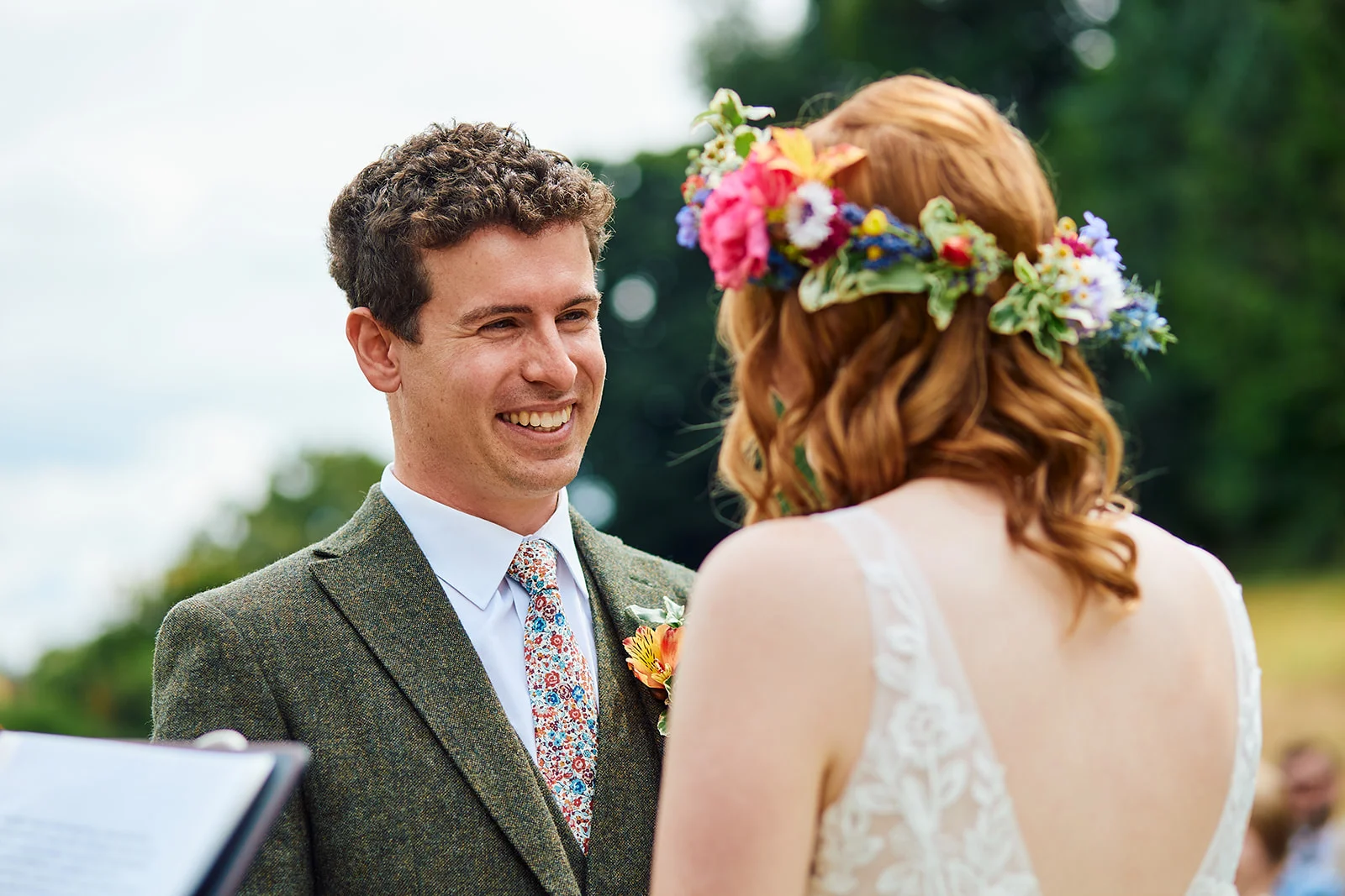 A smiling groom in a tweed suit and floral tie looks lovingly at his bride, who wears a colourful fresh flower crown during an outdoor ceremony.