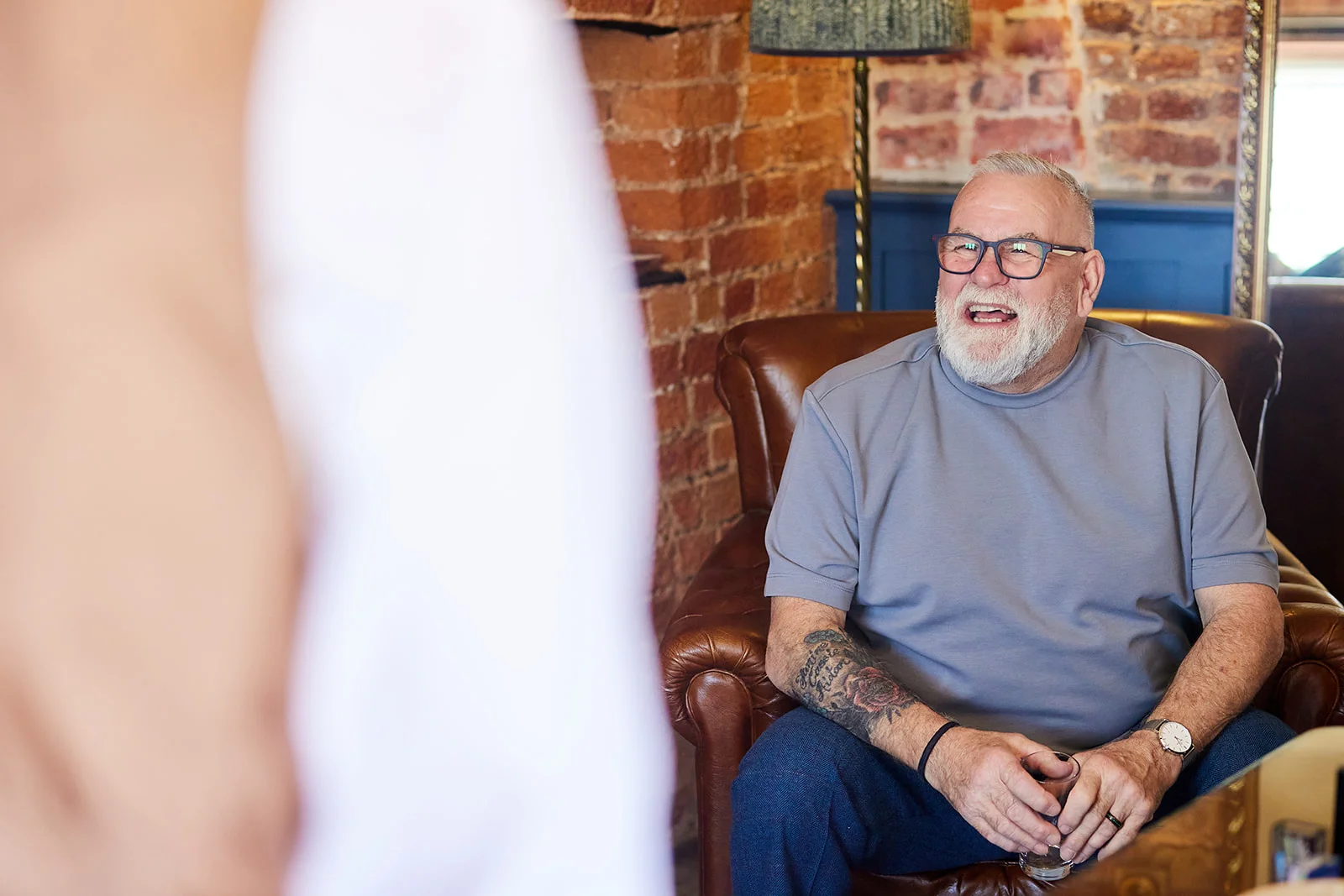 A mature man wearing glasses and a grey t-shirt sits in a leather armchair with exposed brick behind him, laughing as he looks toward someone out of frame.