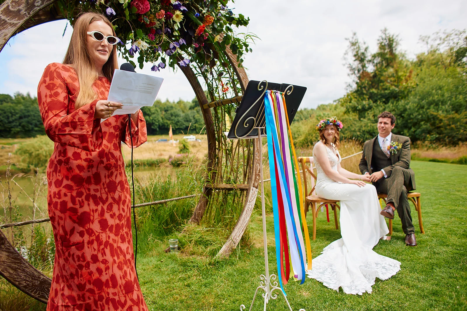 A celebrant wearing a patterned orange dress and white sunglasses reads from notes during an outdoor humanist ceremony, with the bride and groom seated under a floral arch.