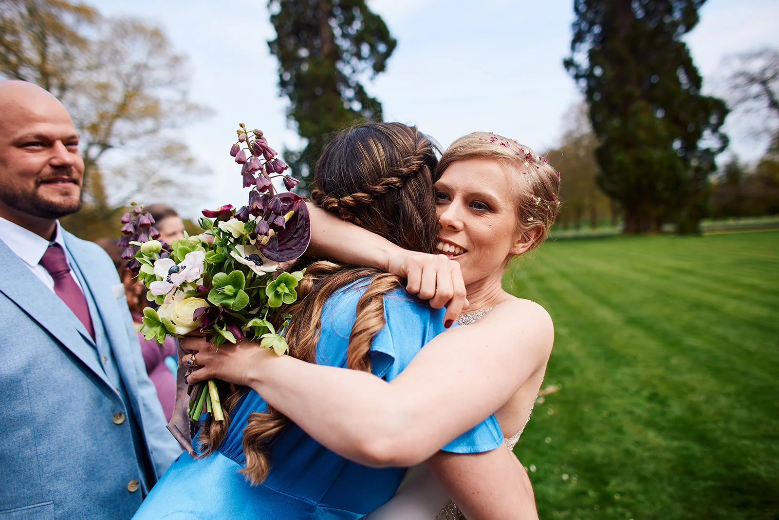 A joyful bride embraces a bridesmaid wearing a vibrant blue dress while holding a wild botanical bouquet outdoors in a lush garden setting.