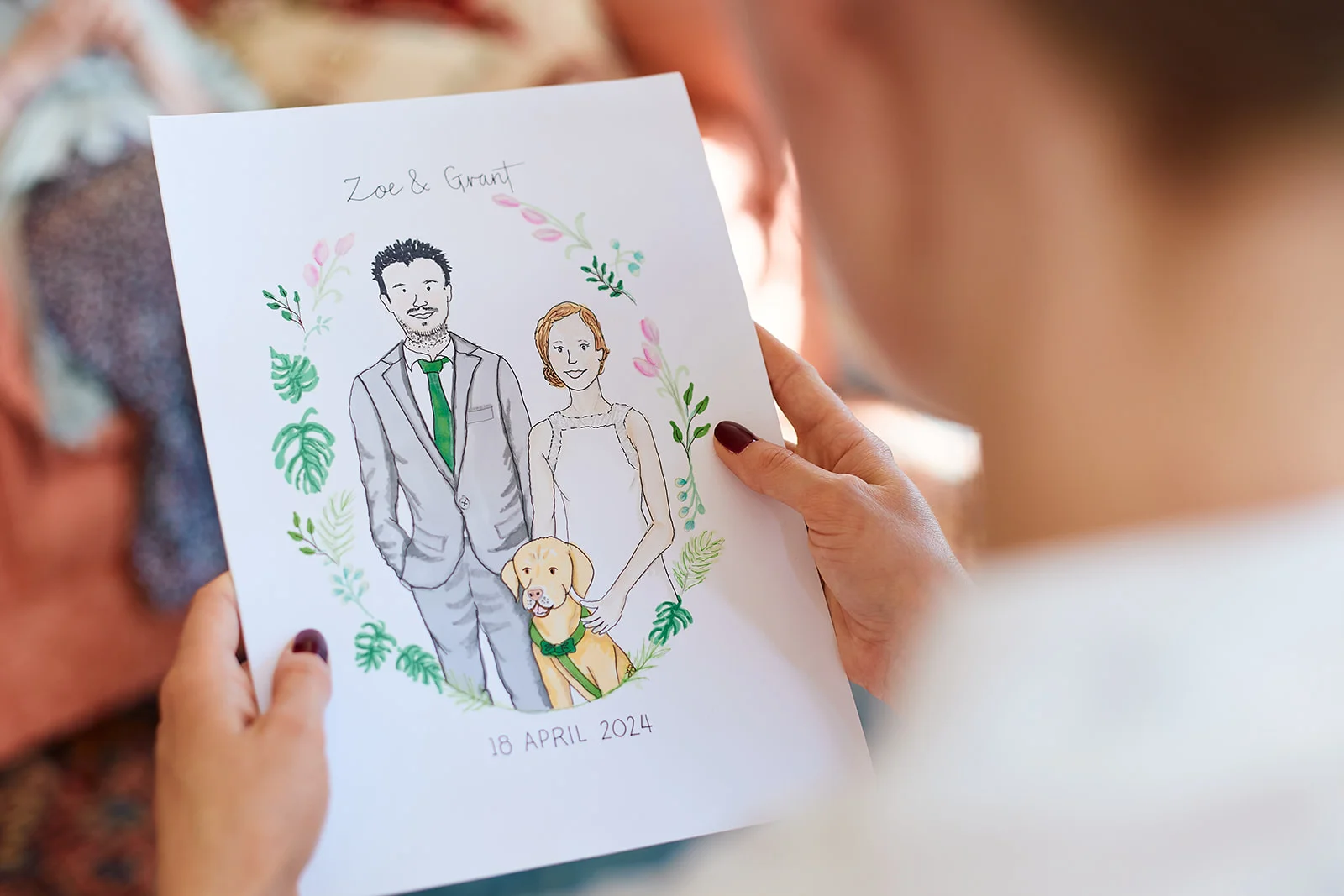 An over the shoulder shot of a person holding a custom watercolour illustration featuring a couple and their dog surrounded by a floral wreath.