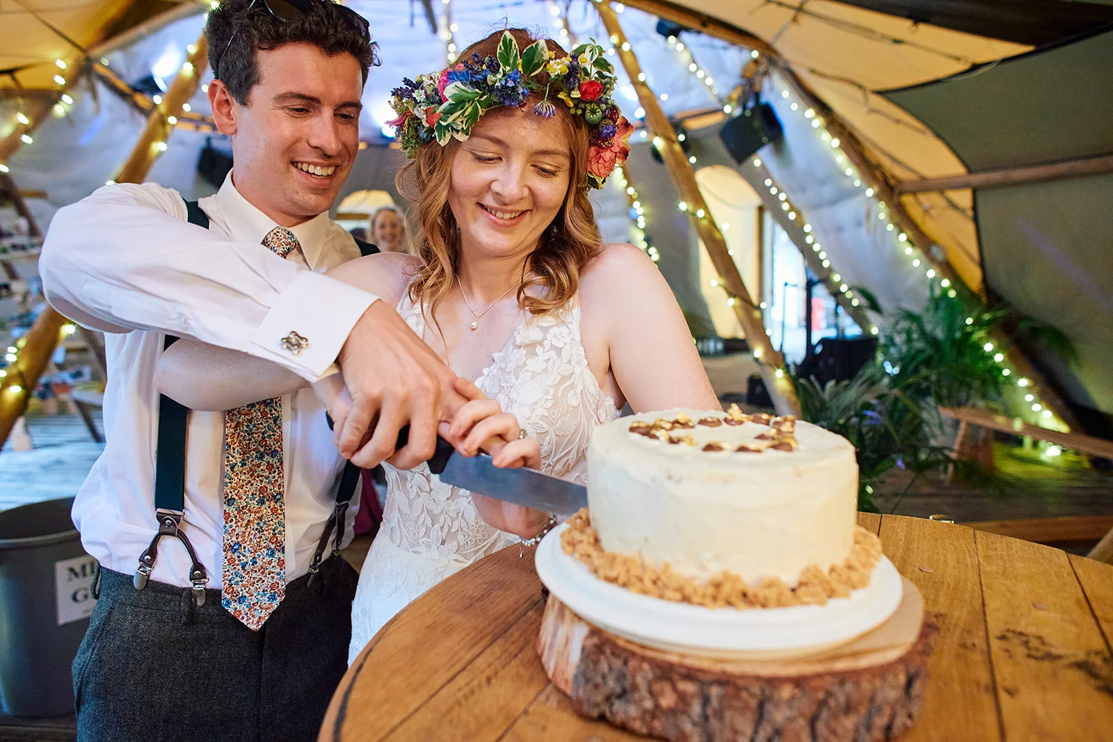 A smiling bride wearing a vibrant floral crown and the groom with braces happily cut their white-frosted wedding cake inside a warmly lit tipi.