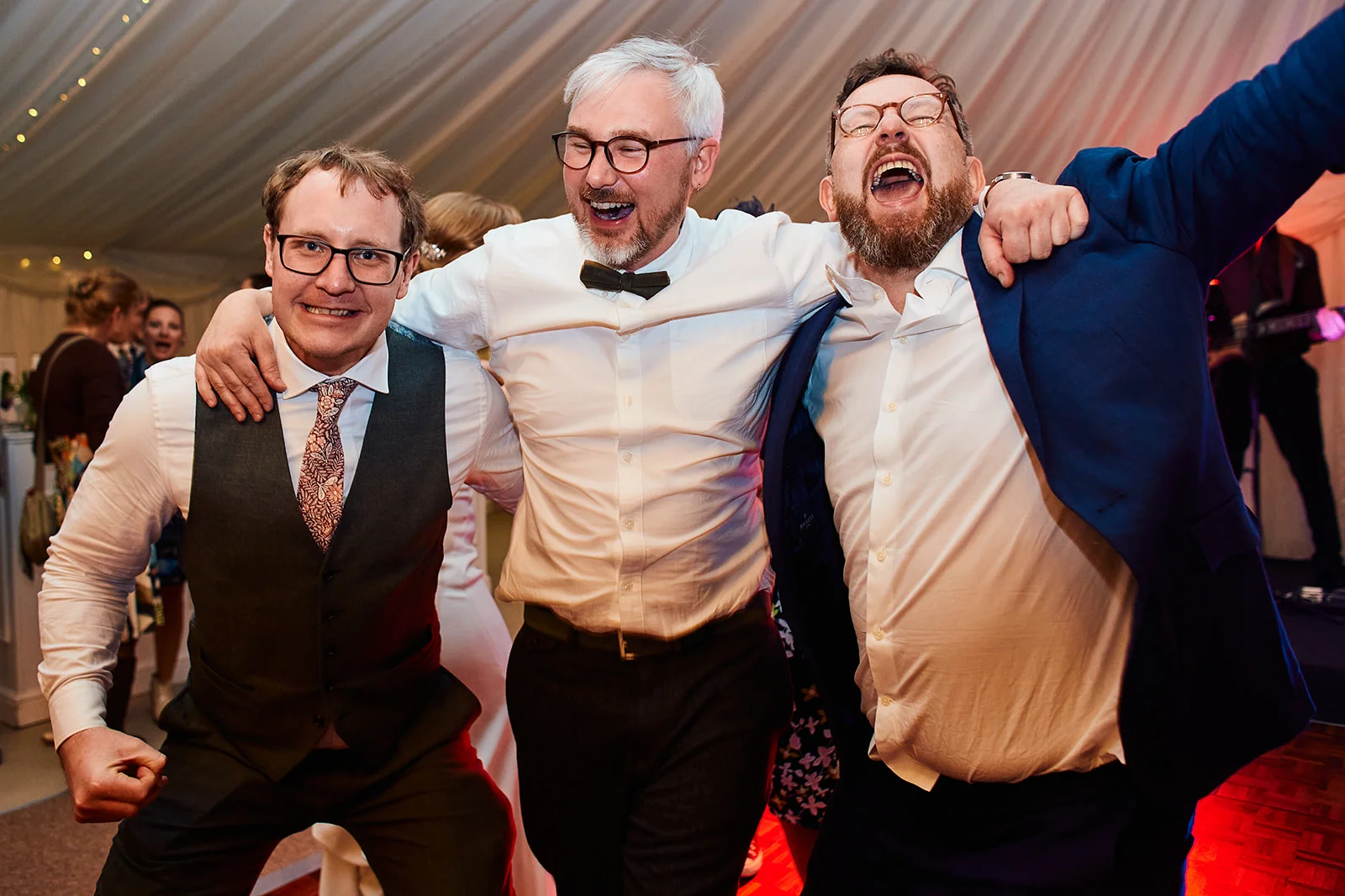 Three smiling men with arms around each other's shoulders enjoying themselves on the dance floor inside a marquee wedding reception.
