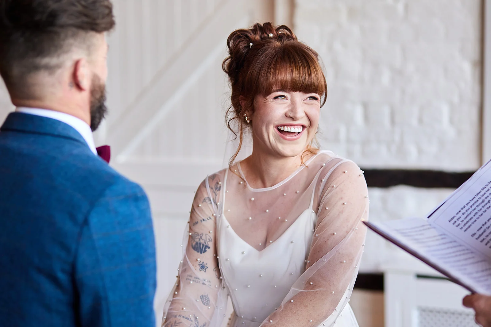 A joyful bride with red hair and tattoos laughs heartily during her wedding ceremony while facing her groom in a blue suit.