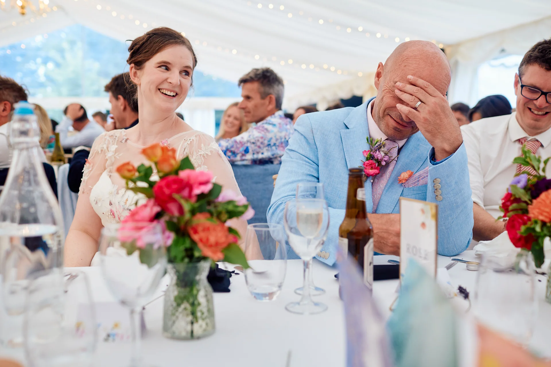 A laughing bride and a groom covering his face with his hand sit at a top table during wedding breakfast speeches inside a brightly lit marquee.