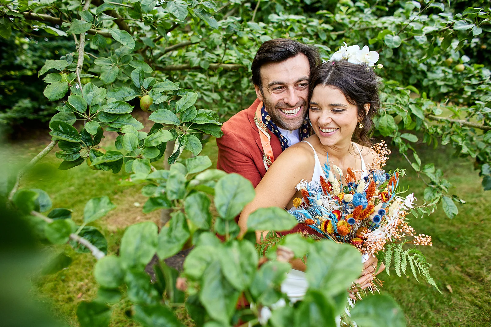 A happy bride and groom embrace joyfully within an apple orchard, framed by green leaves while the bride holds a vibrant dried flower bouquet.