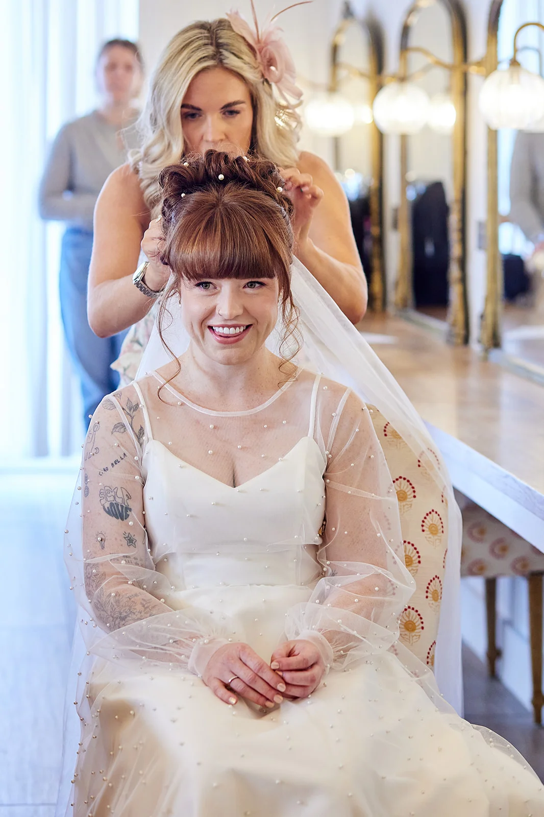 A smiling bride with a fringe and tattoos sits in front of a mirror as a stylist puts the finishing touches to her pearl-adorned bridal updo.