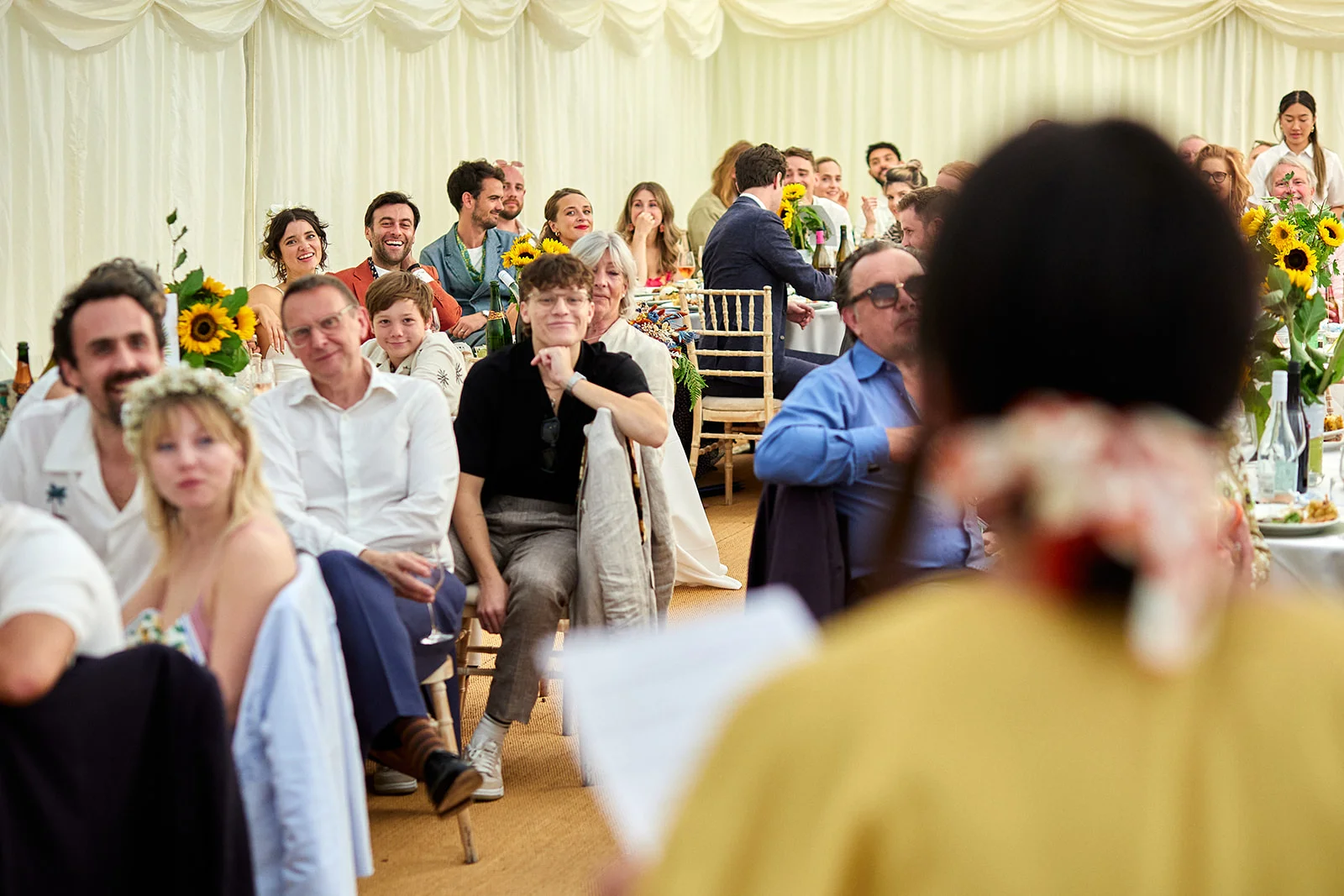 An over the shoulder shot of a speaker delivering a wedding speech inside a brightly lit marquee, with wedding guests listening and laughing during the wedding breakfast.