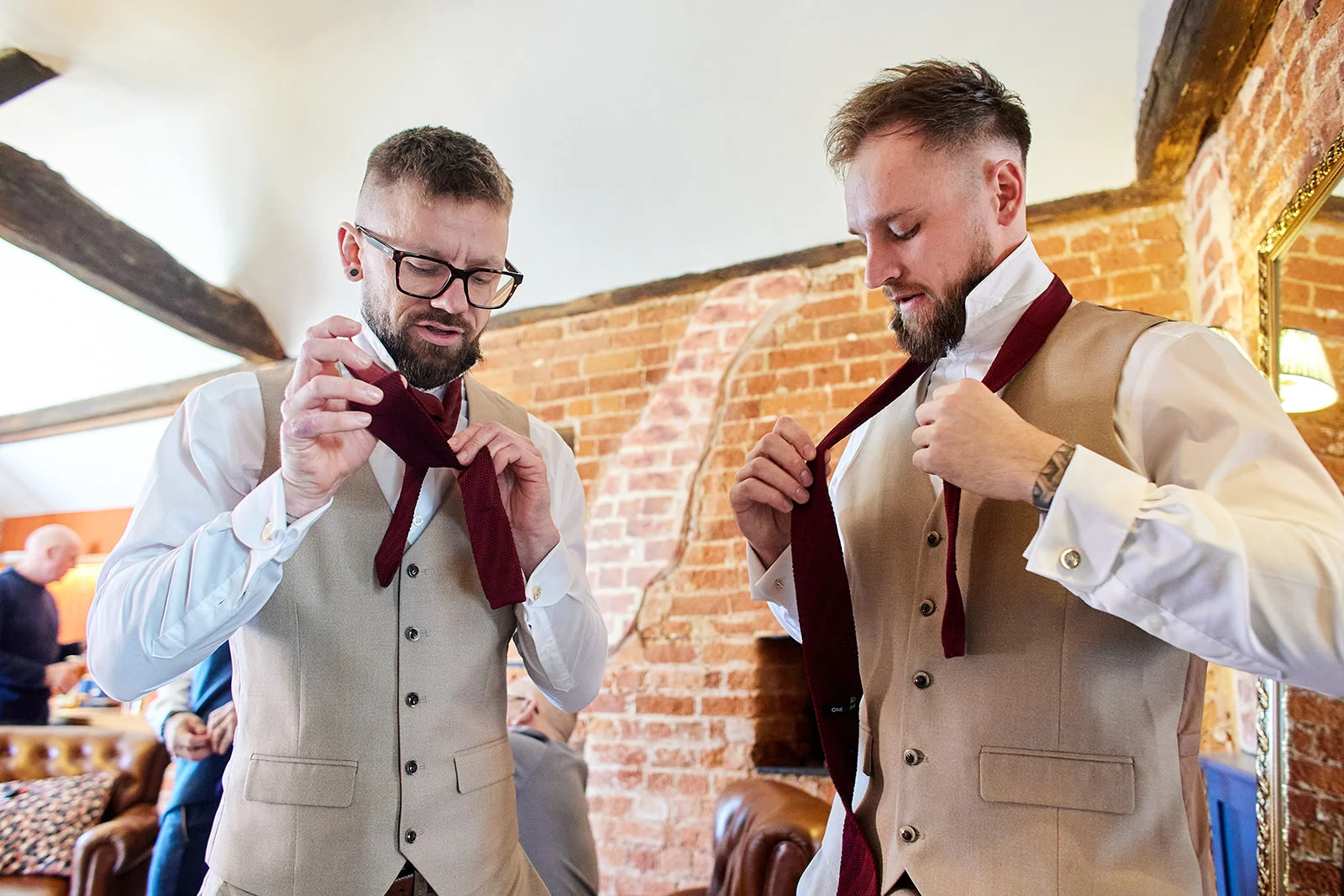 Two grooms getting ready together in a rustic barn venue with exposed brick walls, carefully adjusting their burgundy ties over beige waistcoats.
