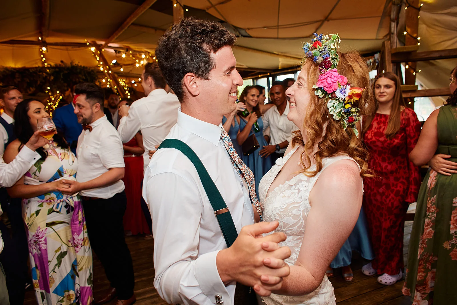 A newlywed couple shares a joyful first dance inside a festoon-lit stretch tent marquee, surrounded by cheering guests.