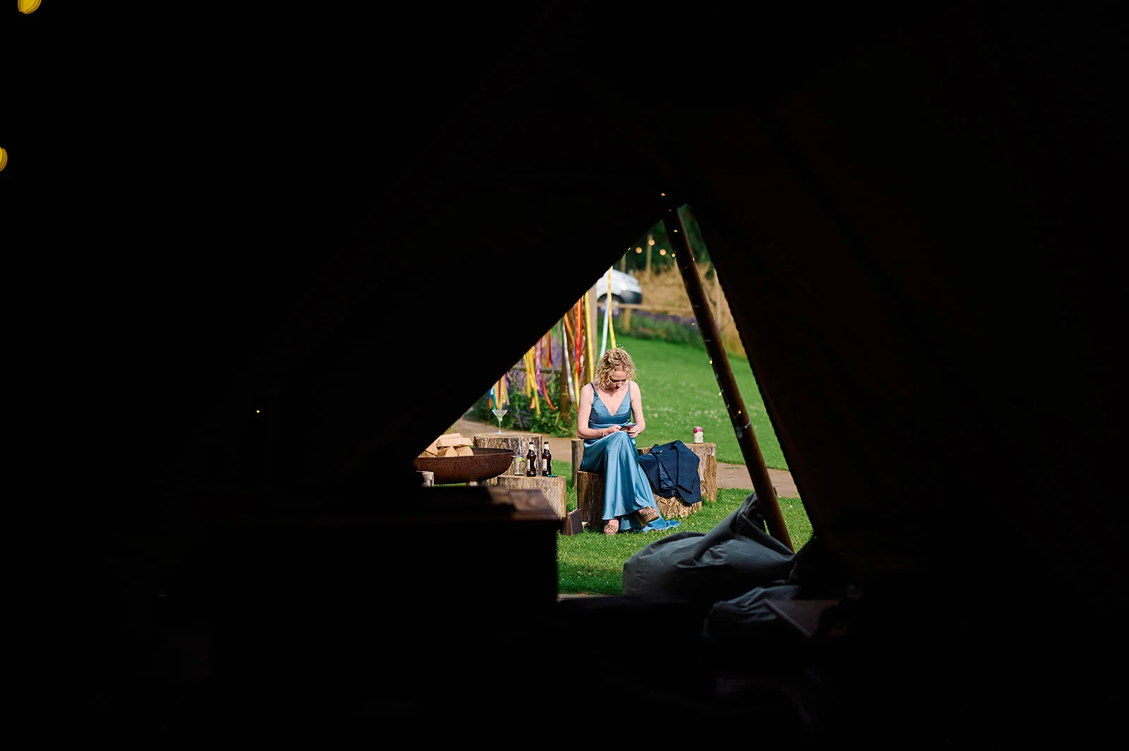 A bridesmaid in a blue dress sits outside on a wooden stump, framed by the dark triangular opening of a tipi tent at a wedding.