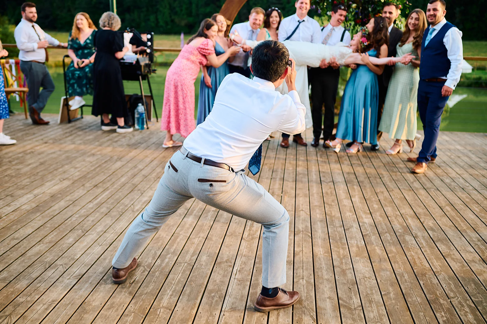 A wedding photographer in a wide stance captures a playful moment as friends lift the bride horizontally on an outdoor wooden deck.
