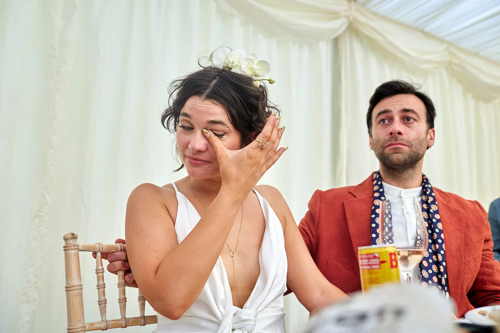 An emotional bride wipes away happy tears while seated at the top table in a draped marquee alongside her partner.
