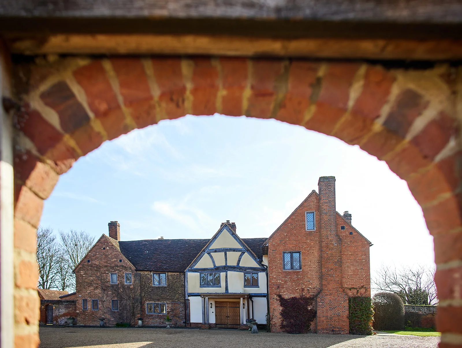 A historic red brick and timber-framed manor house venue is viewed through a traditional brick archway under a bright blue sky.