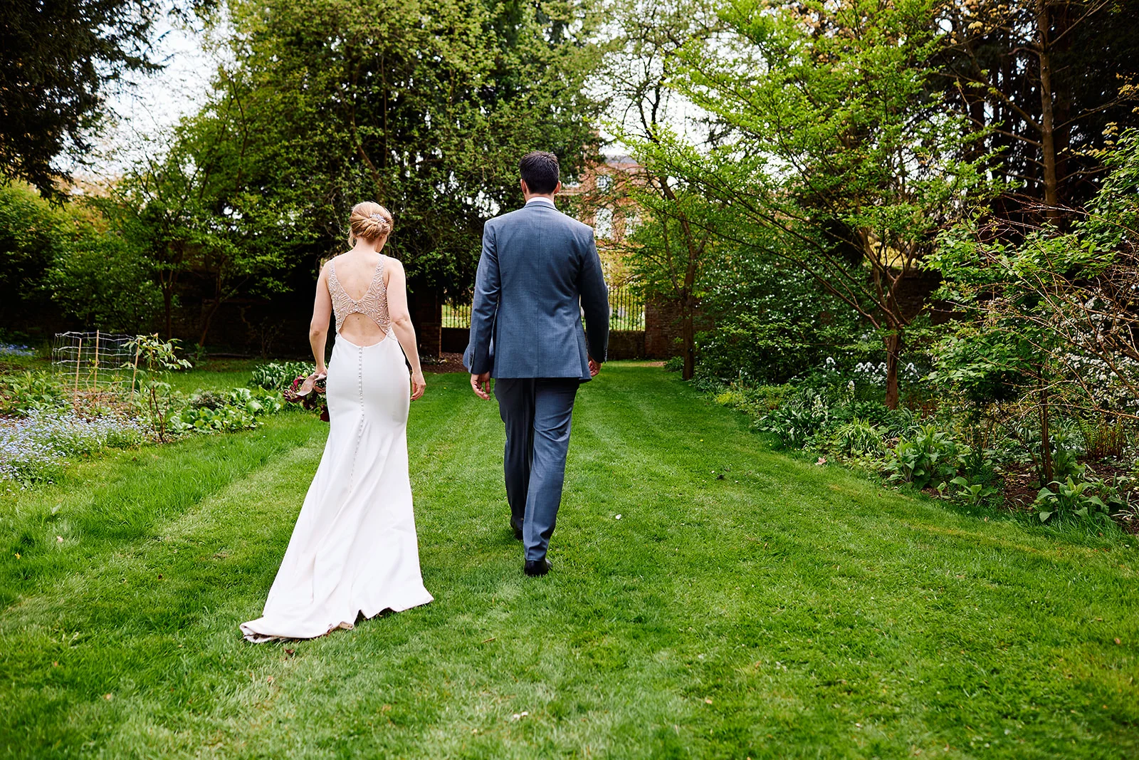 Newlyweds walk away down a lush green lawn in a country house garden, viewed from behind with the bride wearing an open-back crepe wedding gown and the groom in a grey suit.