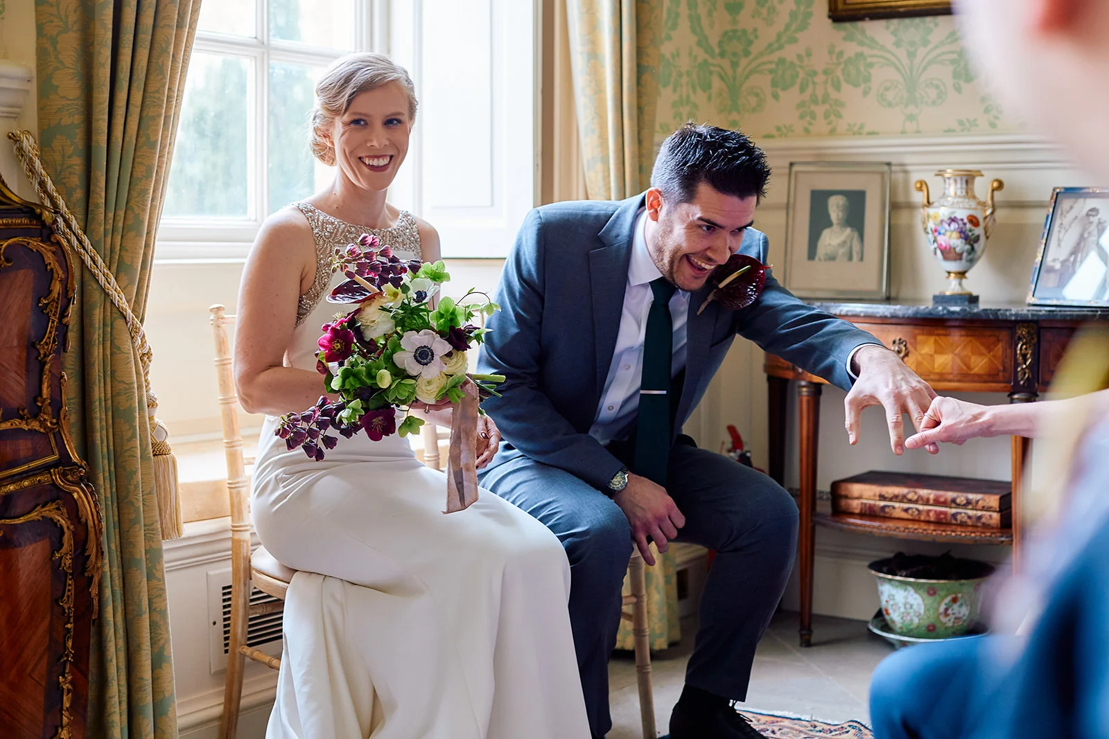 A newly married couple sitting indoors near a bright window share a joyful moment of laughter as the groom proudly displays his wedding ring to a guest.