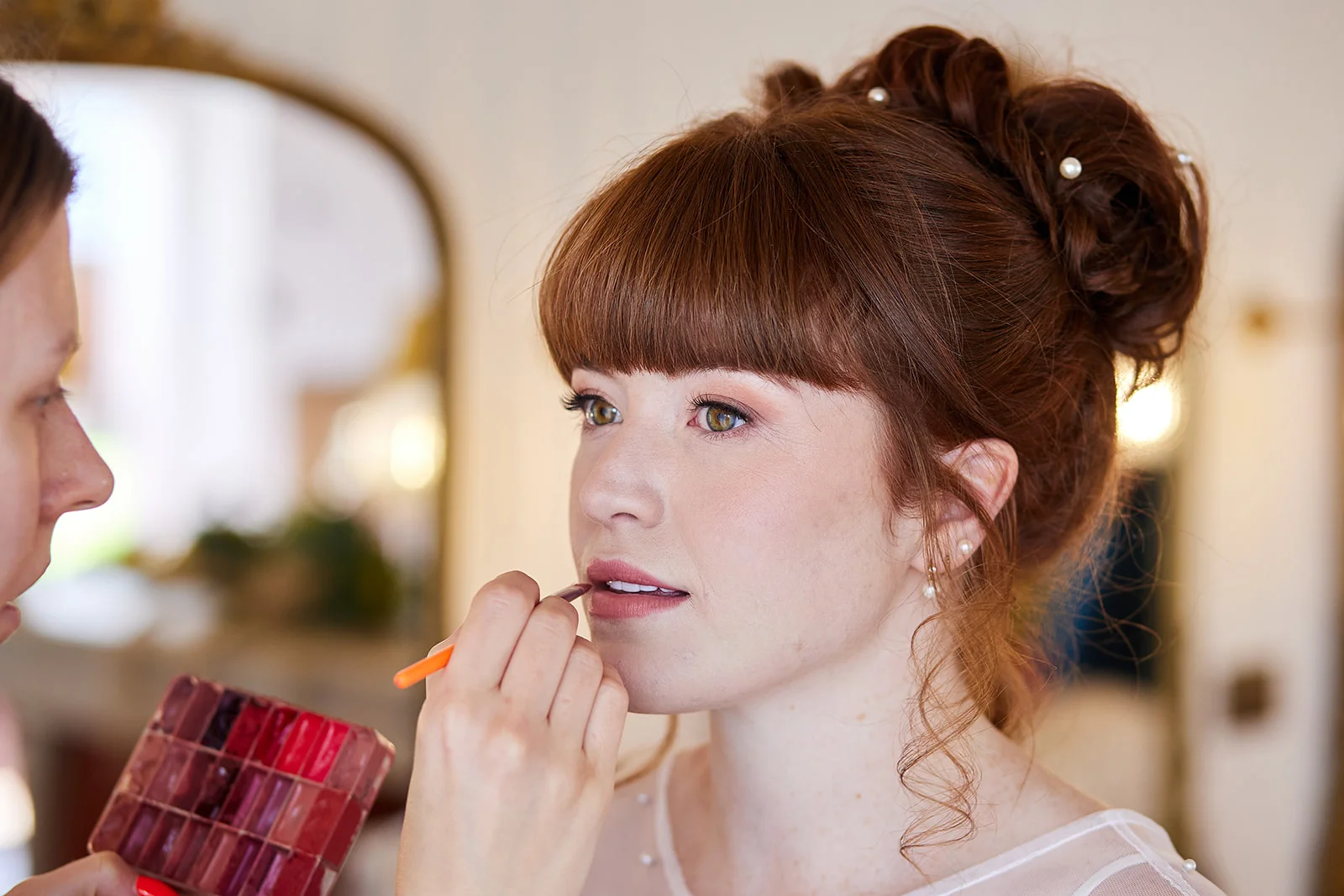 A makeup artist delicately applies lipstick to a bride with auburn hair styled in an updo adorned with pearls during morning preparations.