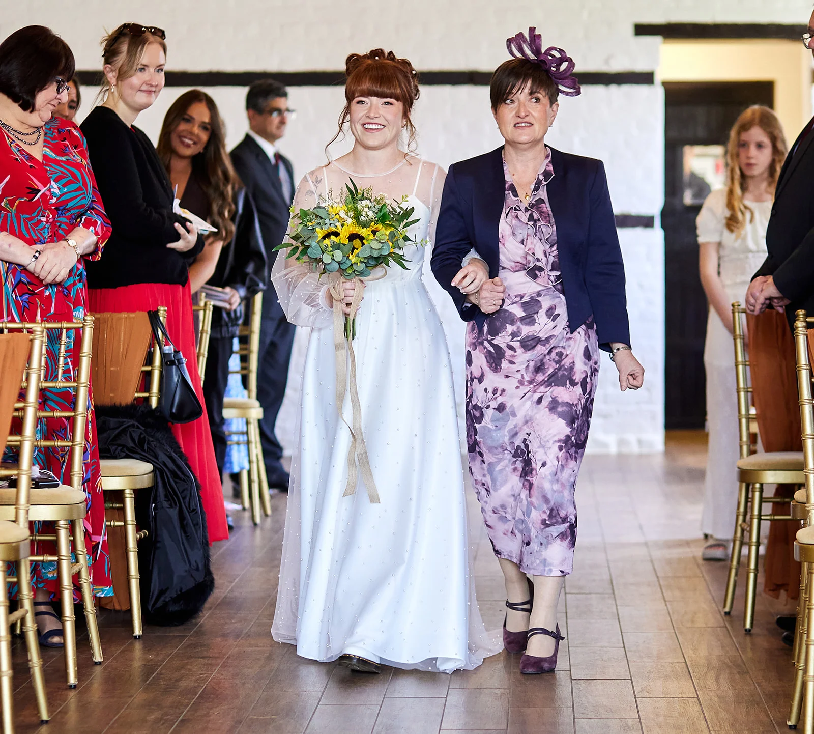 A smiling bride holding a sunflower bouquet walks down the aisle arm-in-arm with her mother, flanked by seated guests watching.