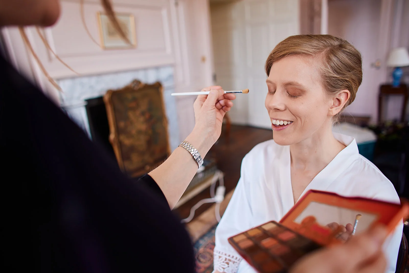 A smiling bride wearing a white getting-ready robe has her eyeshadow applied by a makeup artist inside a historic manor house.