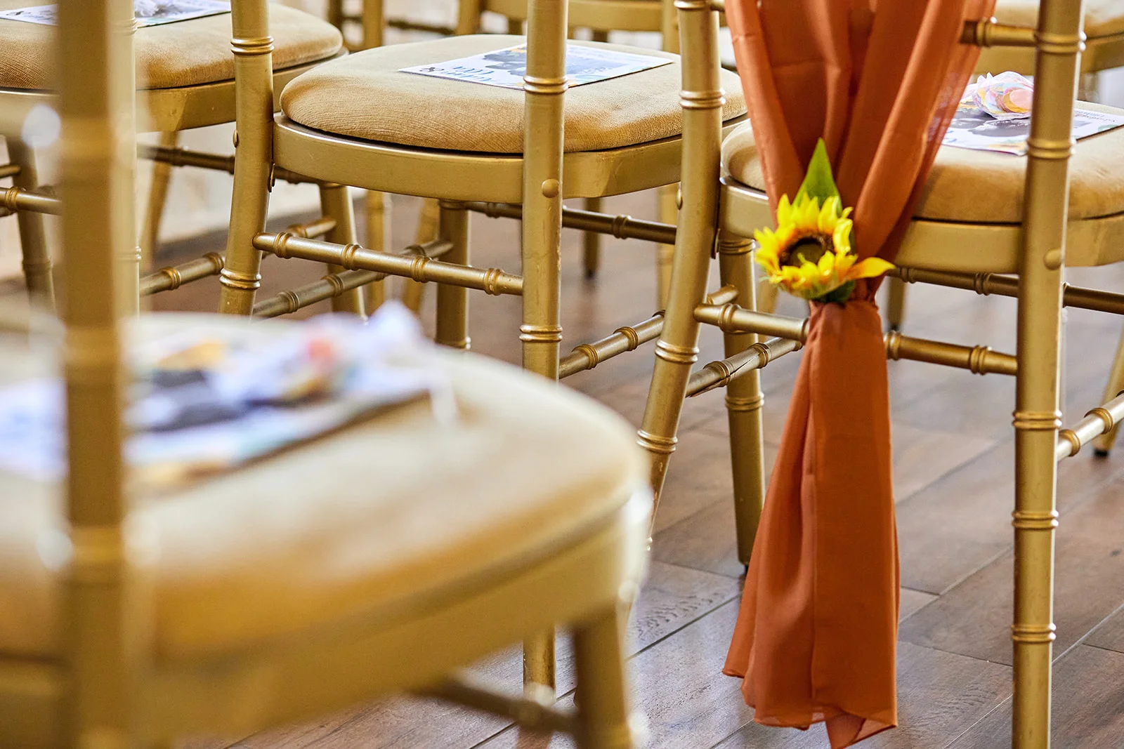 Close up of ceremony chairs adorned with rust-coloured fabric sashes and vibrant sunflower decorations in a light-filled venue.