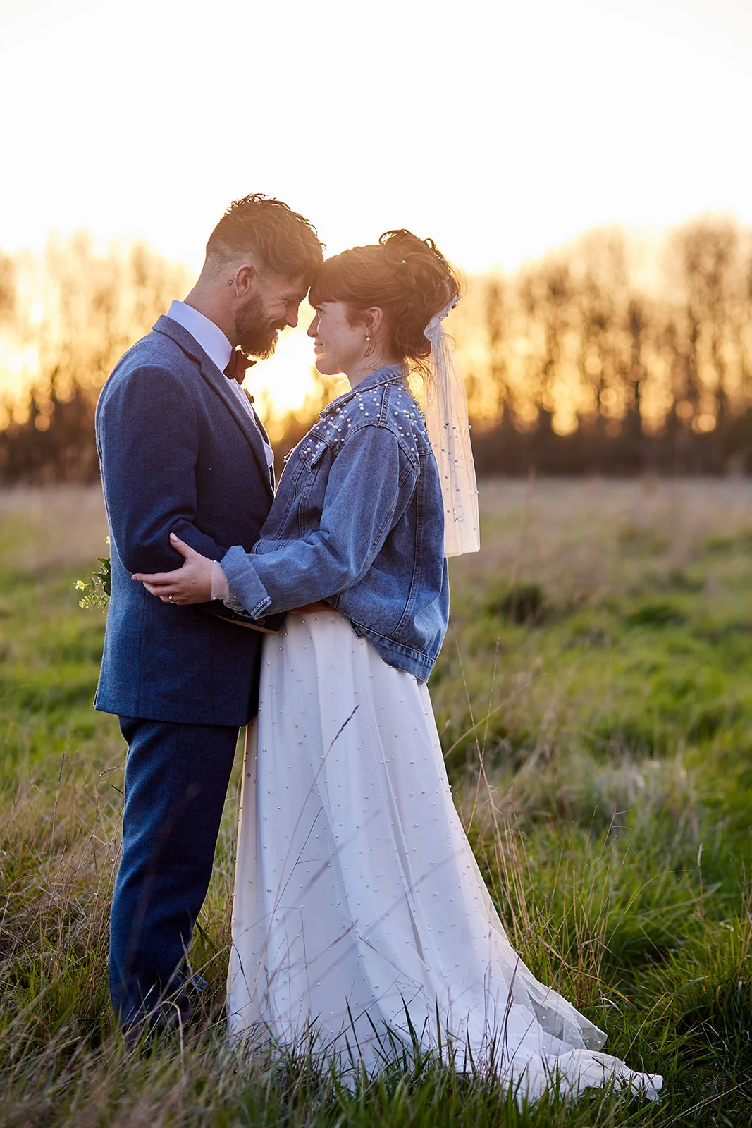 A newlywed couple share a tender forehead kiss in a grassy meadow during golden hour, with the bride wearing a pearl-embellished denim jacket over her gown.