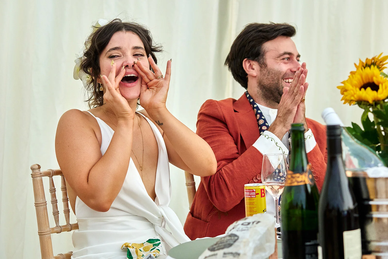 A joyous bride cupping her hands around her laughing mouth alongside a groom in a rust-coloured jacket clapping enthusiastically at the wedding reception.
