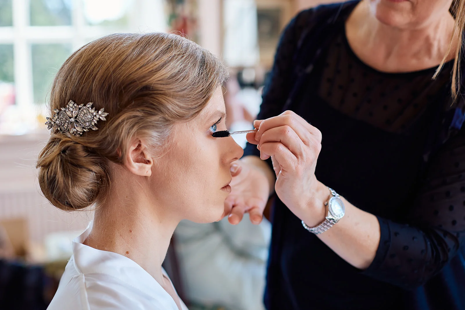 A bride with a low bun and crystal hair comb sits calmly while a makeup artist applies mascara during morning preparations.