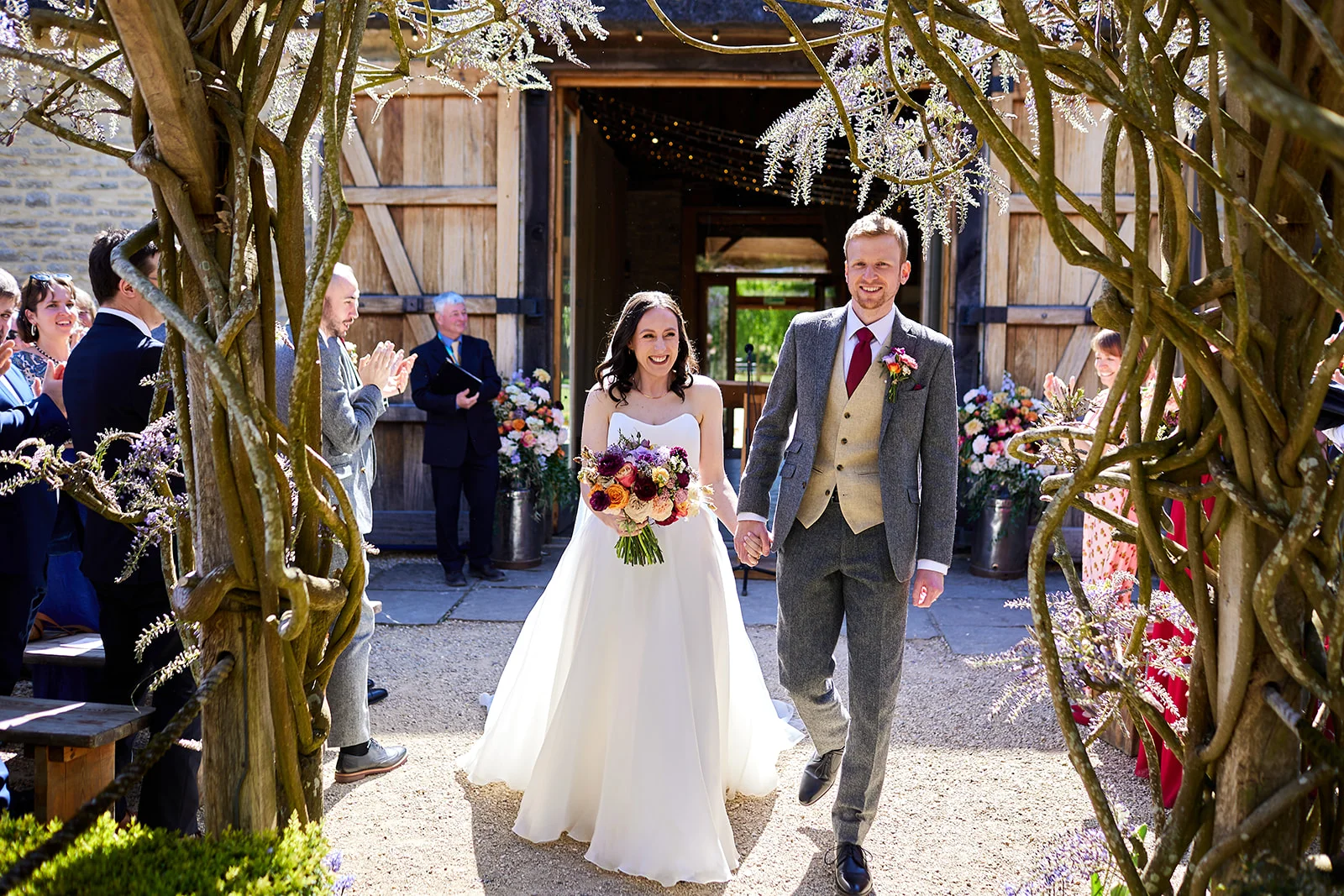 A smiling newlywed couple walks hand in hand out of a rustic stone barn beneath a wisteria-covered archway, cheered on by wedding guests.