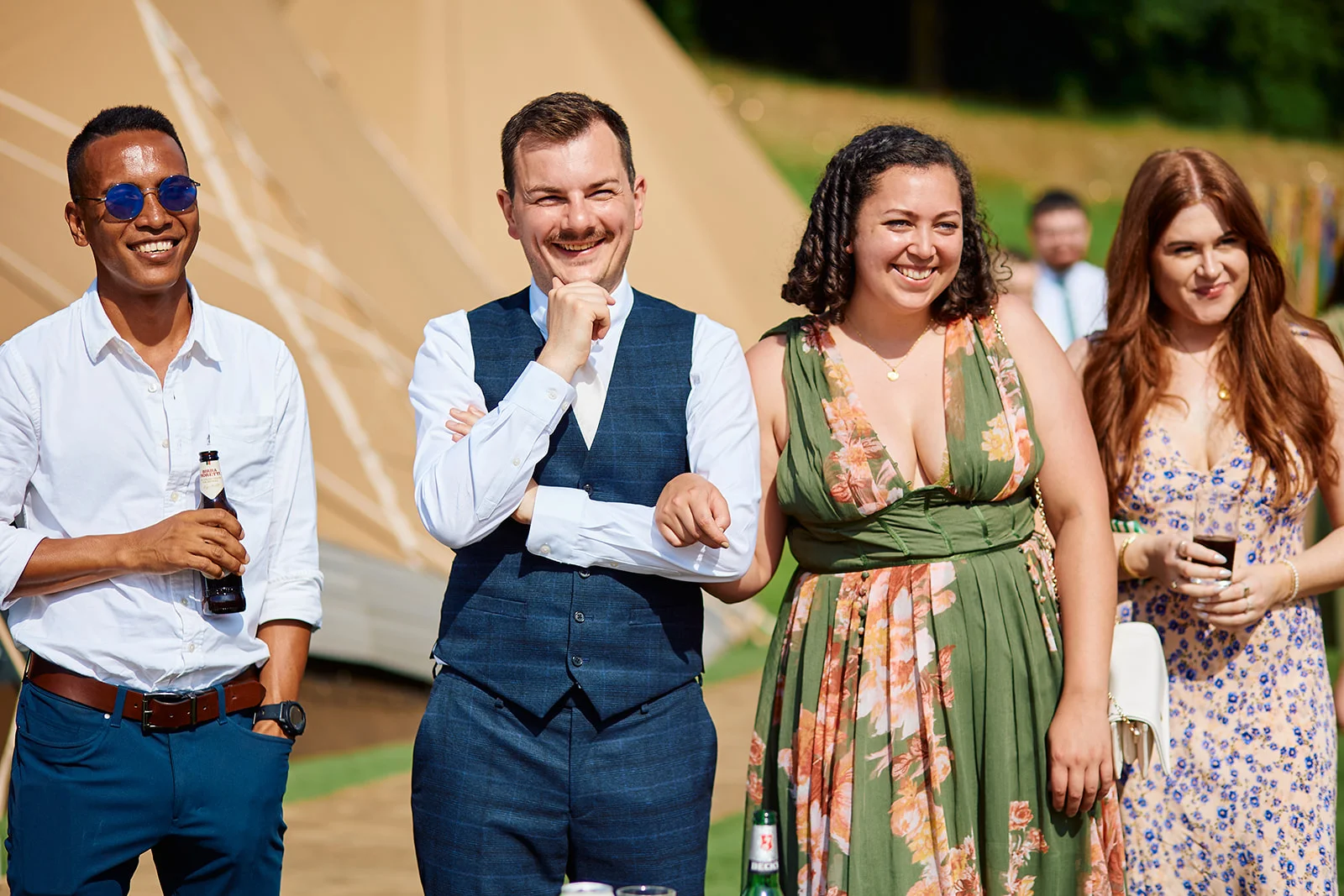 A group of wedding guests smile and mingle outside near a large tipi tent on a sunny day.