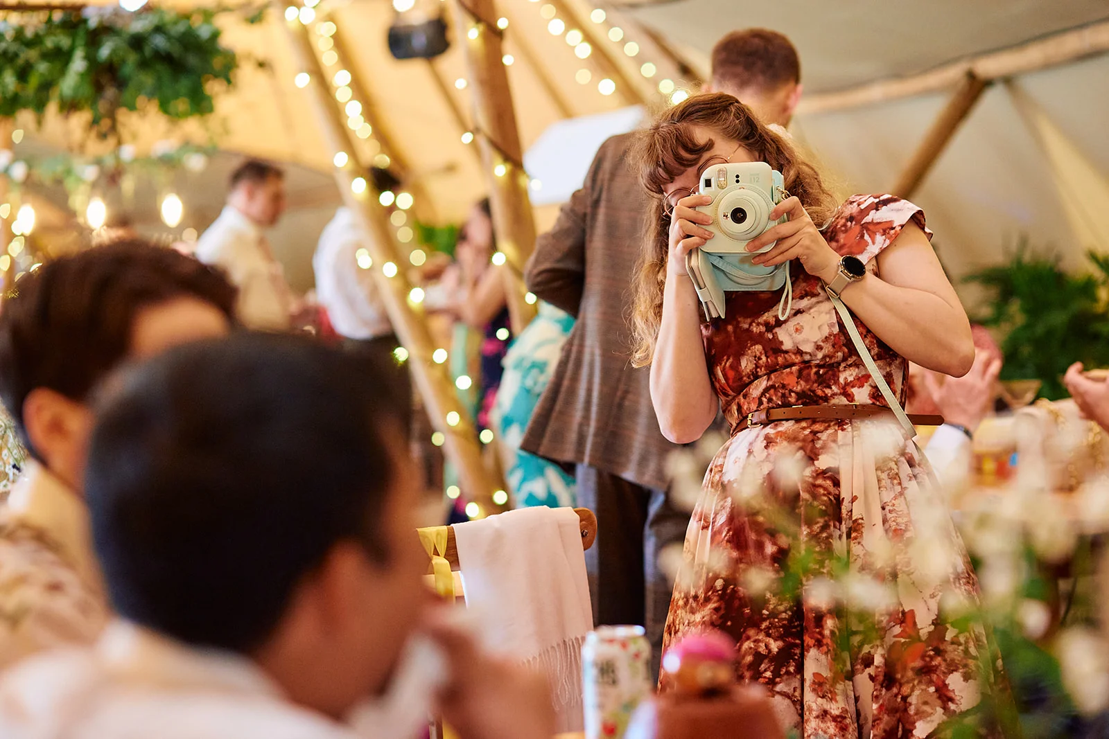A wedding guest taking a photograph with an instant camera inside a festoon-lit tipi marquee during the reception.