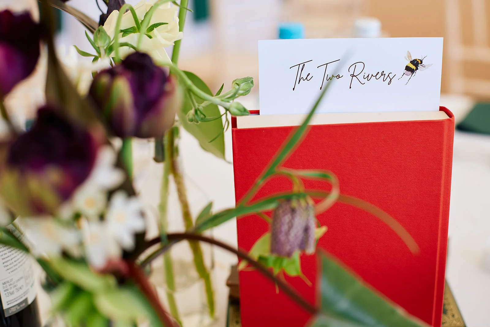 A creative wedding table name card featuring a bee illustration tucked inside a vibrant red book, surrounded by dark purple tulips and delicate wildflowers in bud vases.