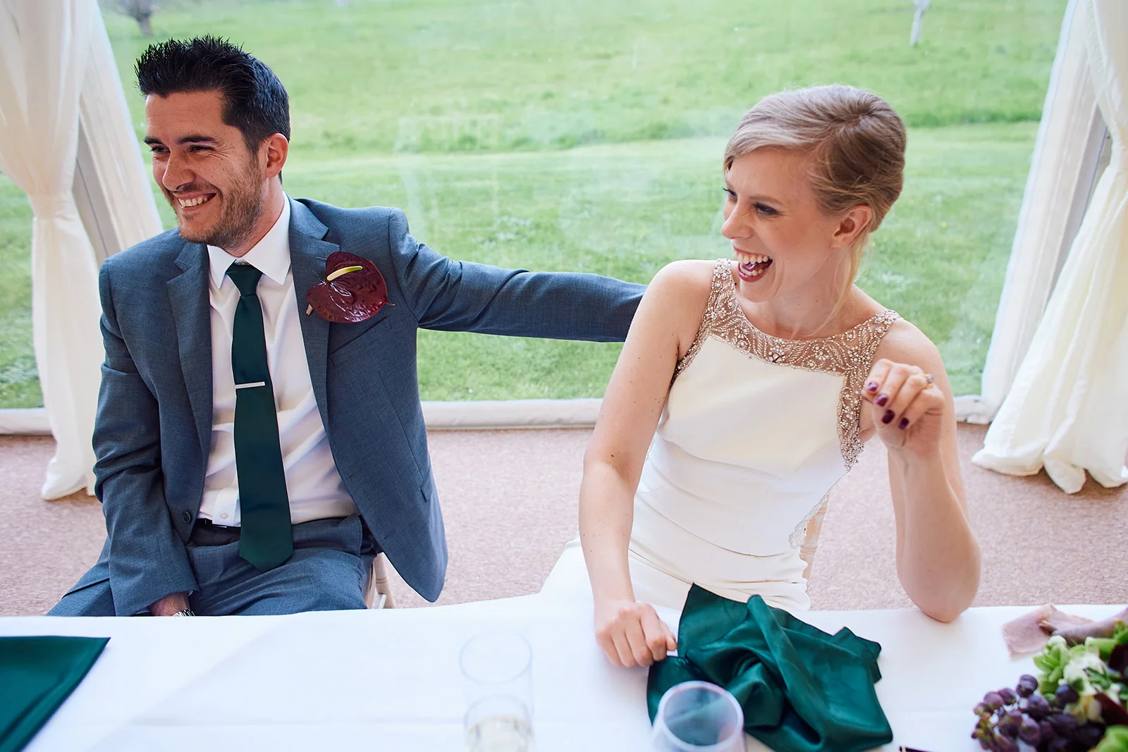 A newlywed couple sitting at the top table in a marquee share a joyful moment of laughter during the wedding speeches.