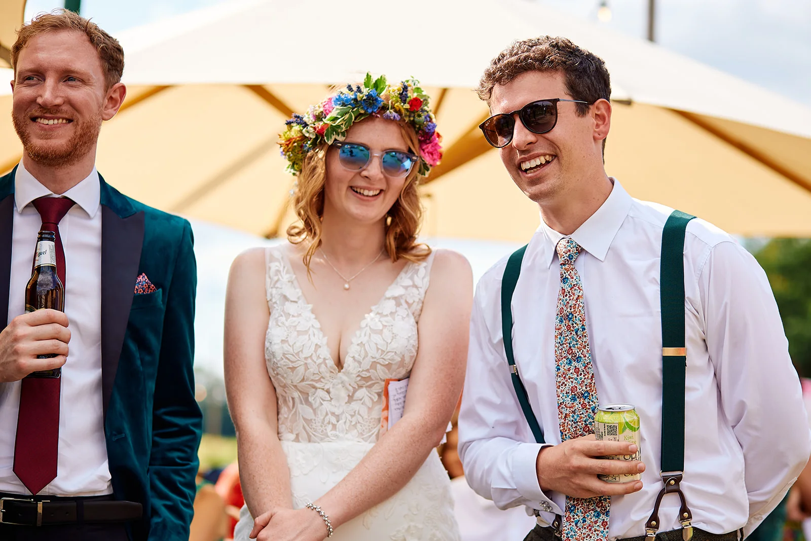 A smiling bride wearing a floral crown and sunglasses stands between two guests holding drinks during a sunny outdoor wedding reception.