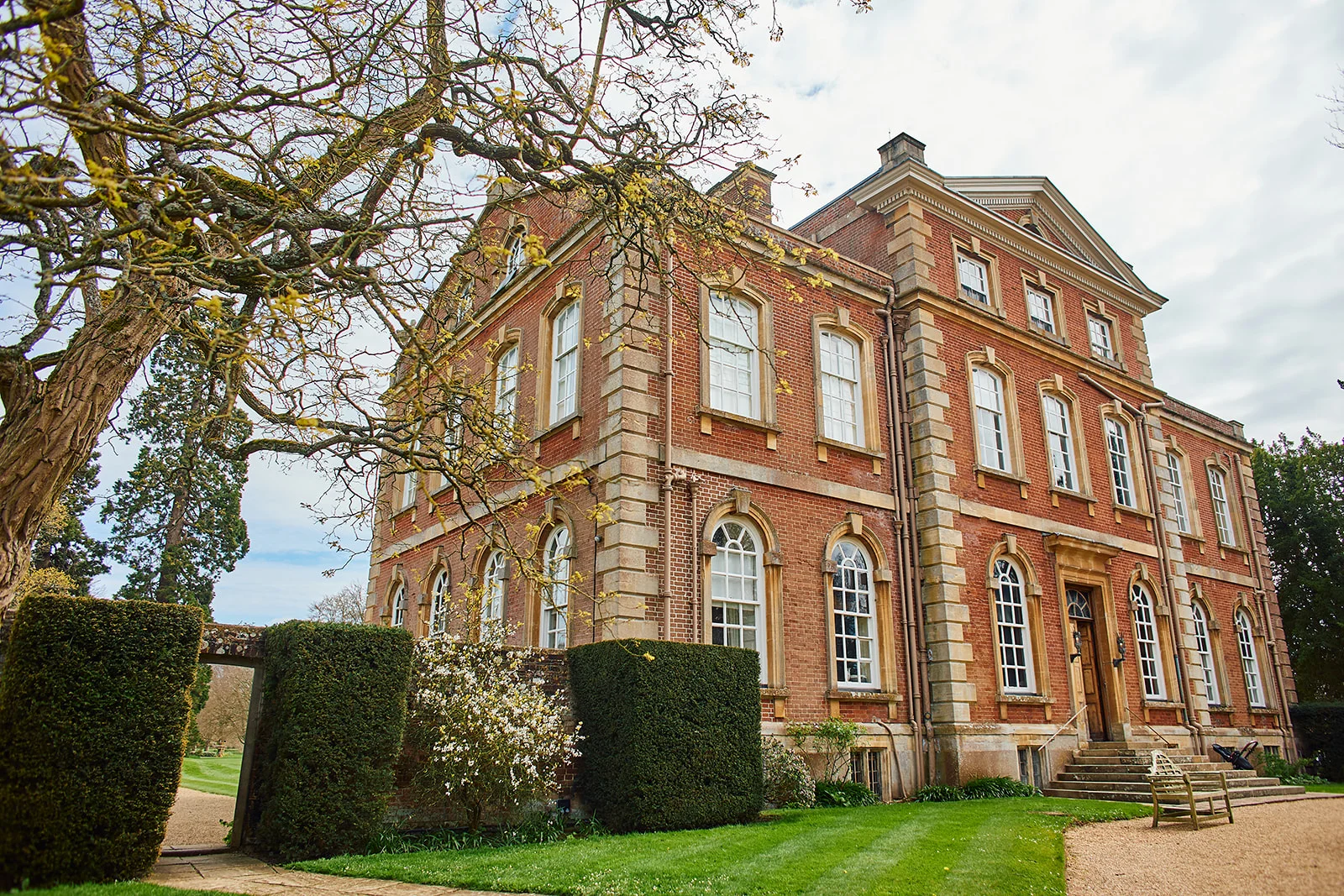 A grand brick country manor house stands against a bright sky, framed by manicured lawns and mature trees during spring.