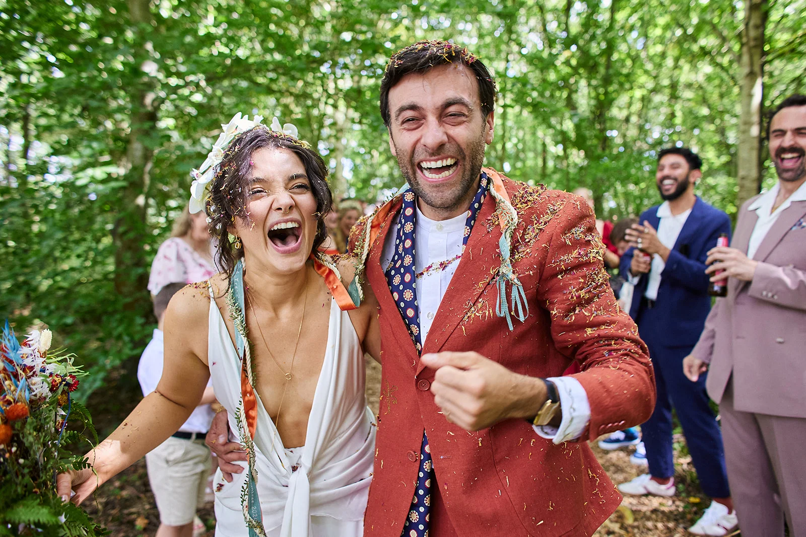 A joyous newlywed couple laughs exuberantly as colorful confetti rains down on them amidst a sunlit woodland wedding celebration.