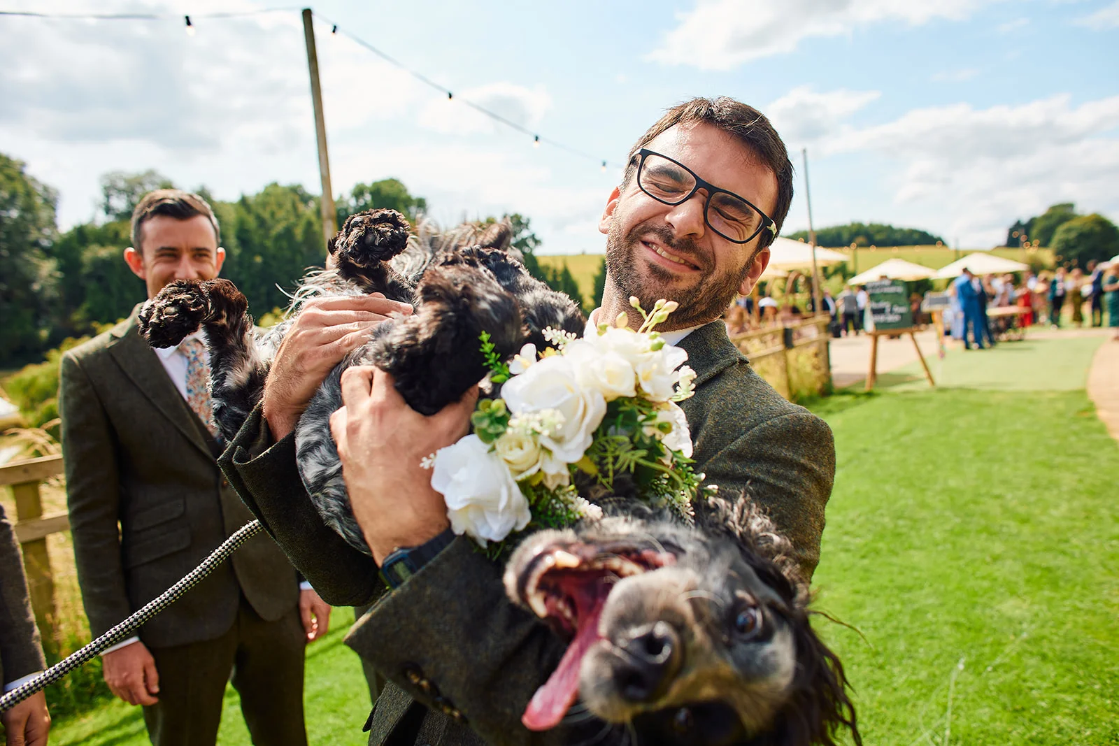 A smiling groom wearing glasses and a tweed suit joyfully holds up a playful dog wearing a floral collar during an outdoor wedding celebration.