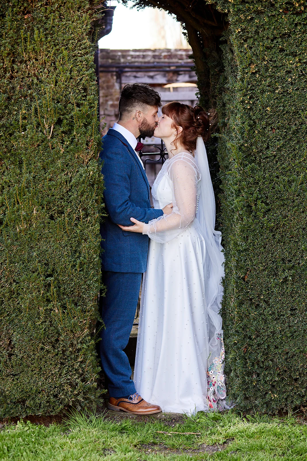 A newlywed couple shares a tender kiss while framed by a shaped topiary arch in an English garden.