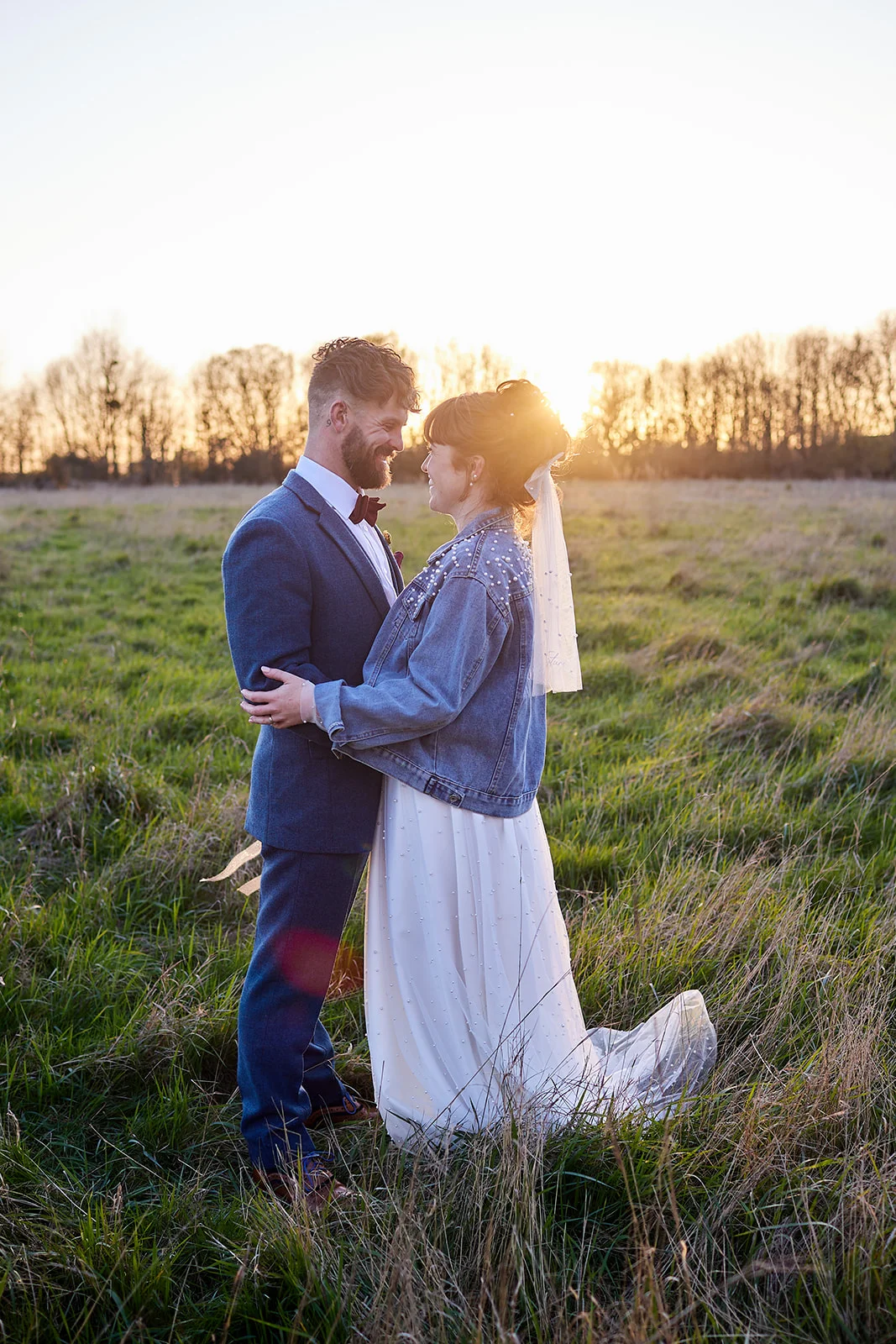 A newlywed couple embraces lovingly in a grassy meadow during sunset, with the bride wearing a pearl-embellished denim jacket over her gown and a bow in her hair.