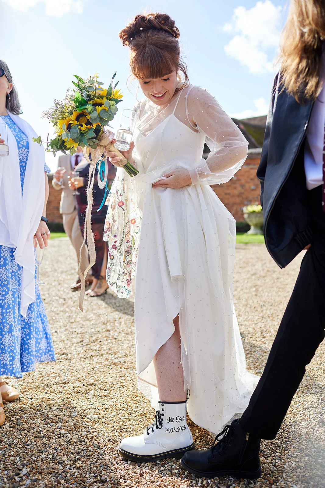 A smiling bride holding a sunflower bouquet and wearing personalized white boots lifts her pearl-embellished gown on a sunlit gravel path.