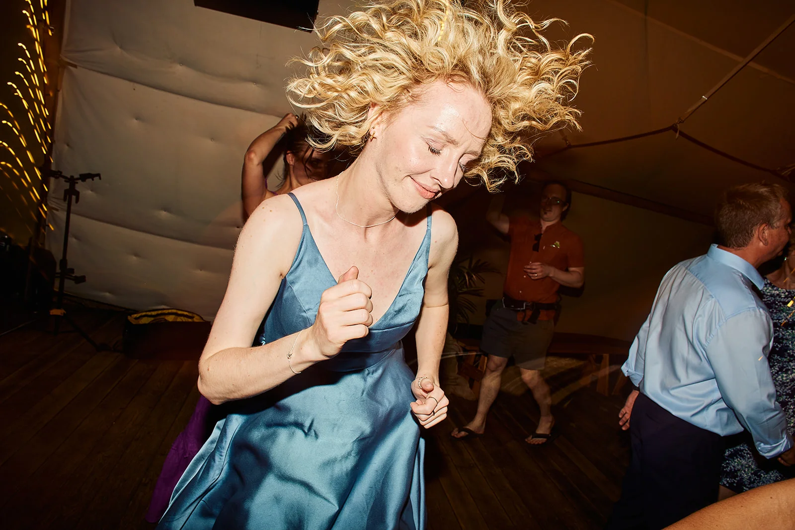 A blonde guest with curly hair tosses her head in joy while dancing vigorously on a crowded evening dance floor.