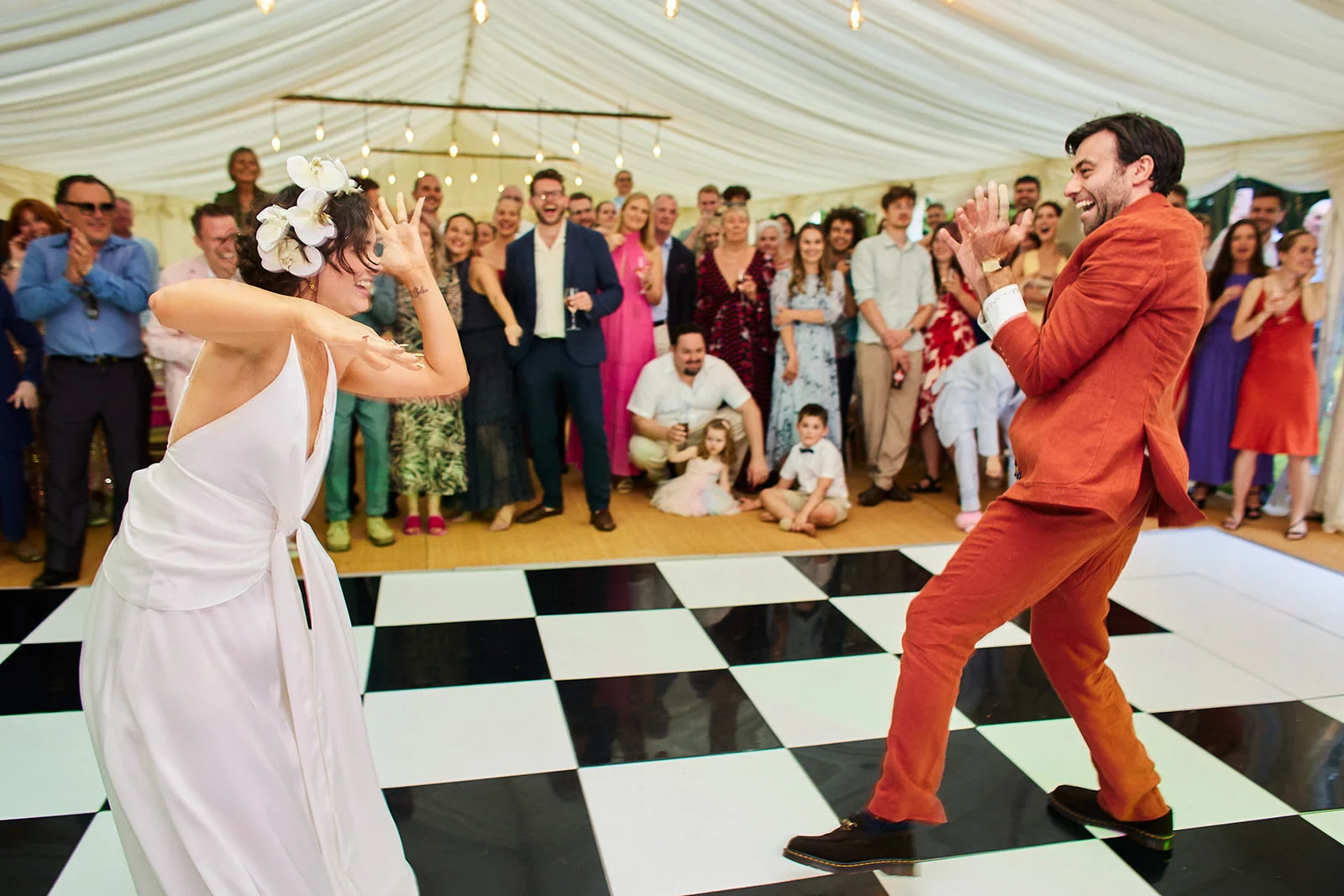 A joyful newlywed couple enthusiastically dance together on a black and white checkerboard dance floor inside a marquee while guests watch and cheer.