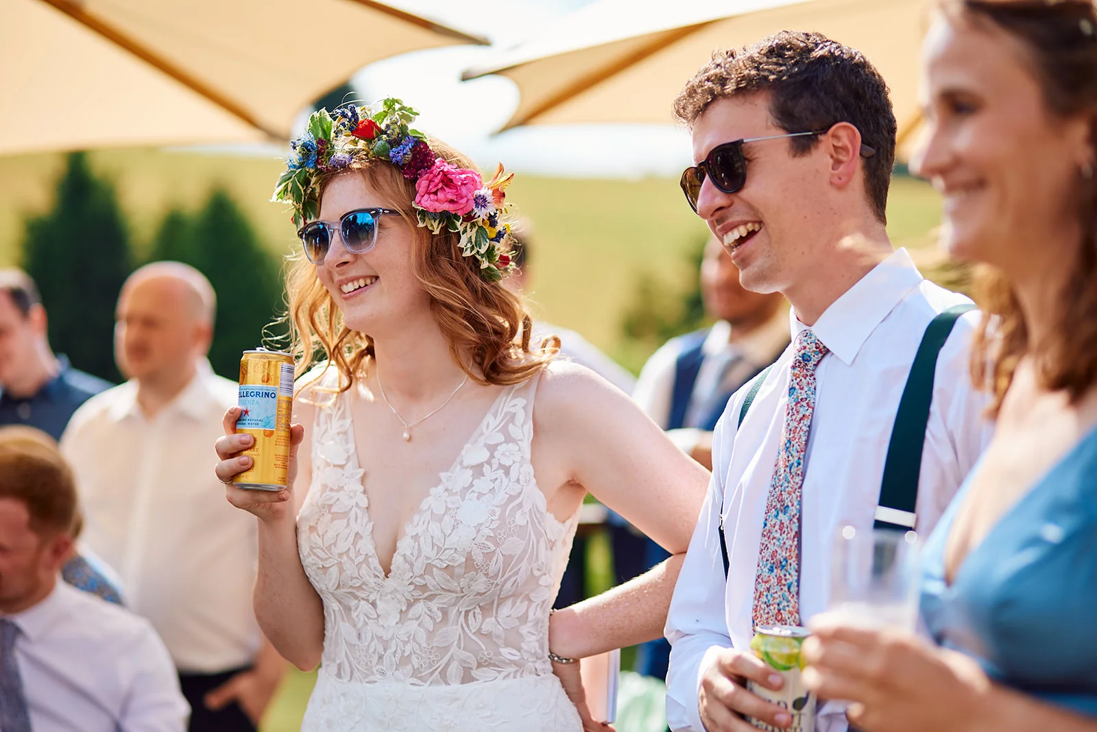 A smiling bride wearing a vibrant floral crown and lace gown enjoys a sunny drinks reception outdoors, holding a can alongside guests.