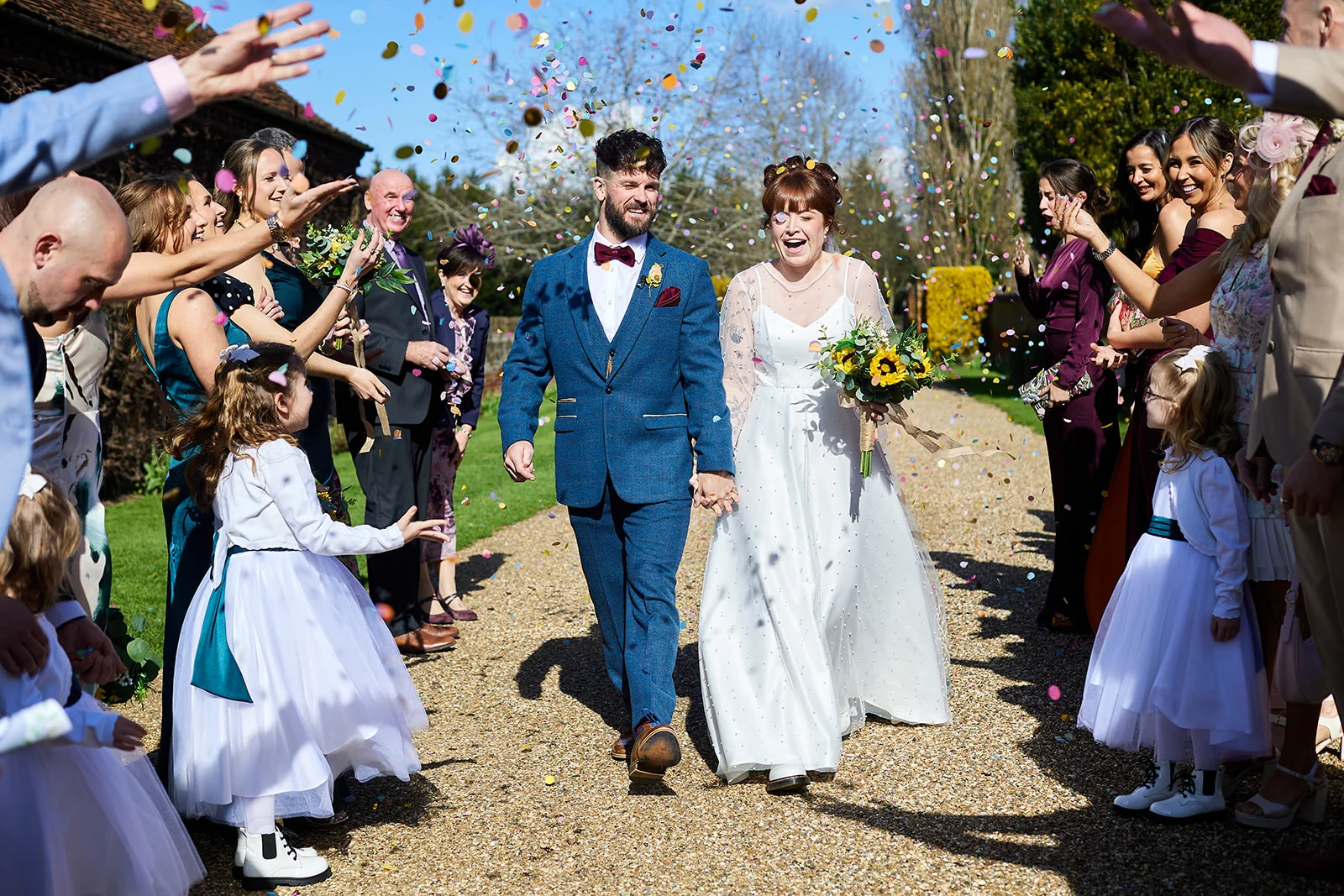 A joyful newlywed couple walks hand in hand down a gravel path while family and friends enthusiastically shower them with colorful confetti.