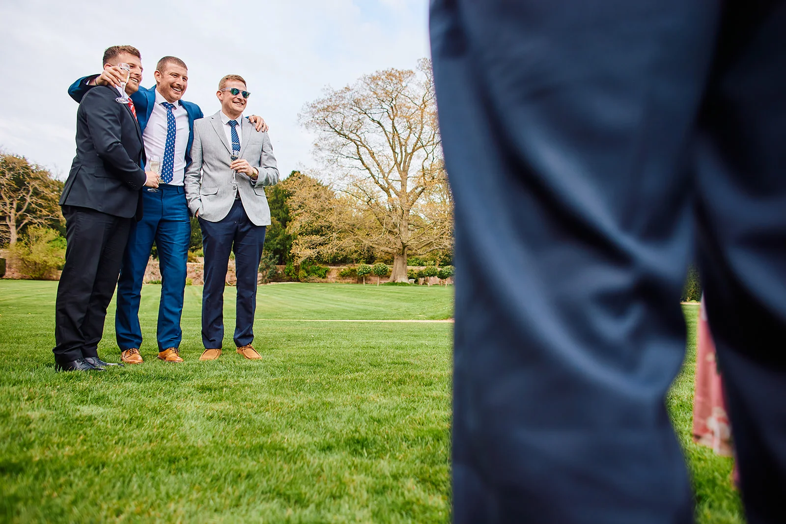 A group of groomsmen stand on a manicured lawn outdoors during a drinks reception, holding glasses and laughing while another guest is blurred in the foreground.