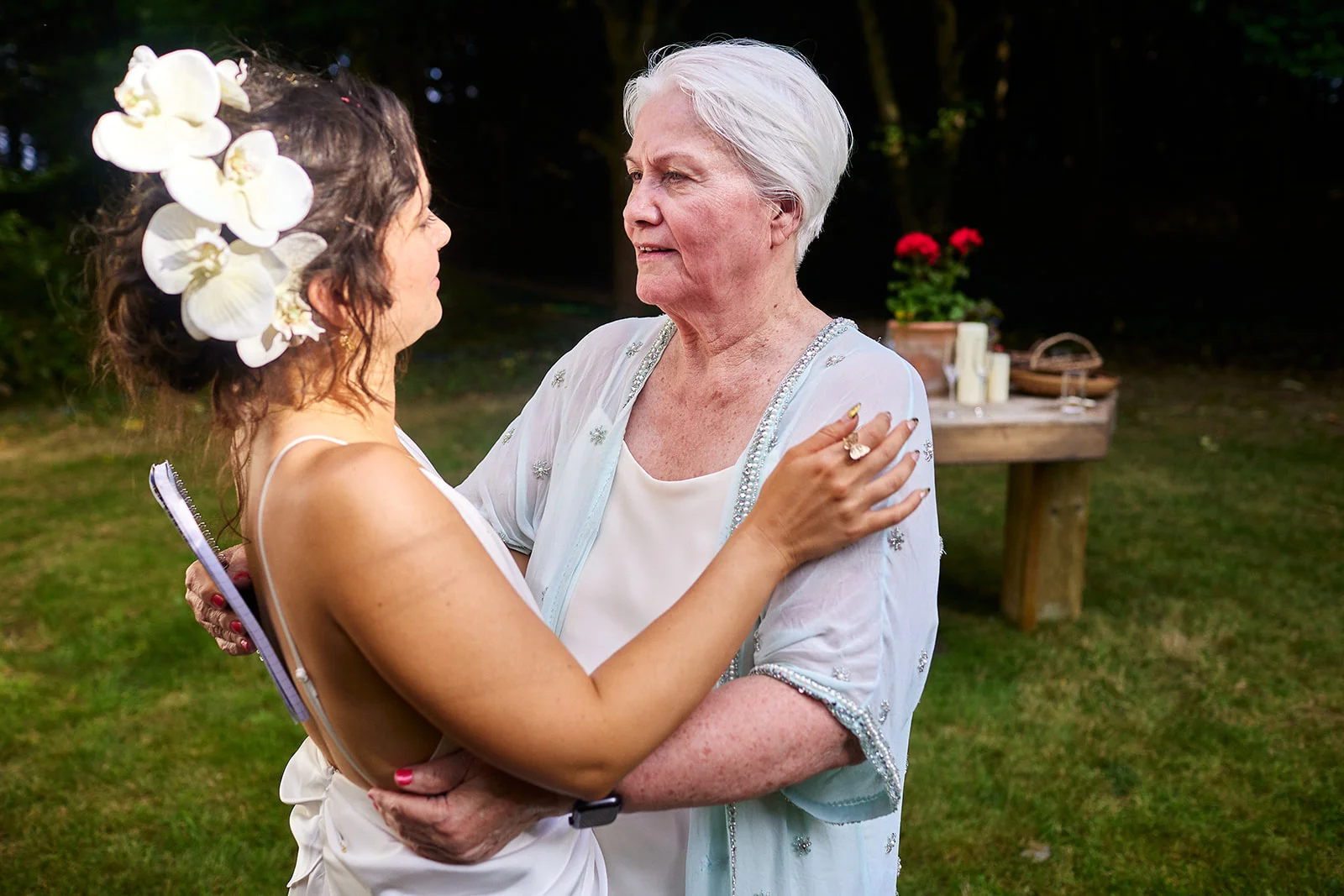 An emotional bride wearing white with white orchids in her hair shares a tender moment outdoors with her mother during the wedding celebration.