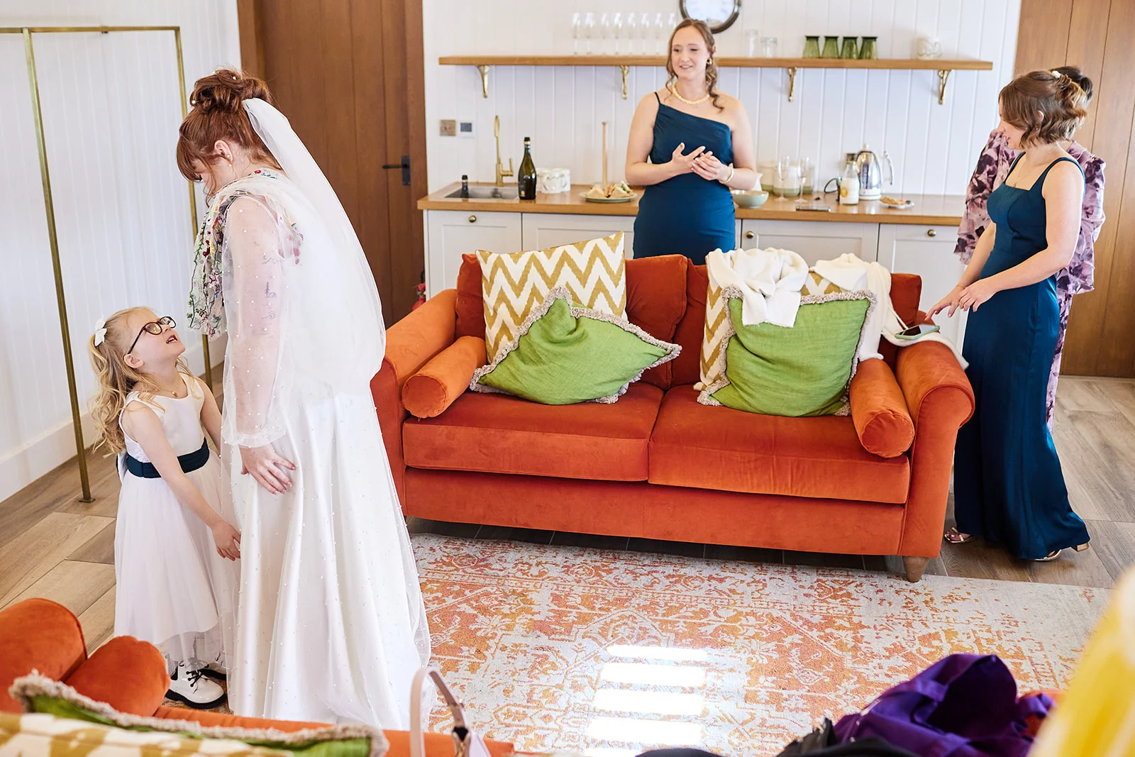 A bride in a white gown and veil stands talking to a flower girl during morning preparations, while bridesmaids watch from a bright room.