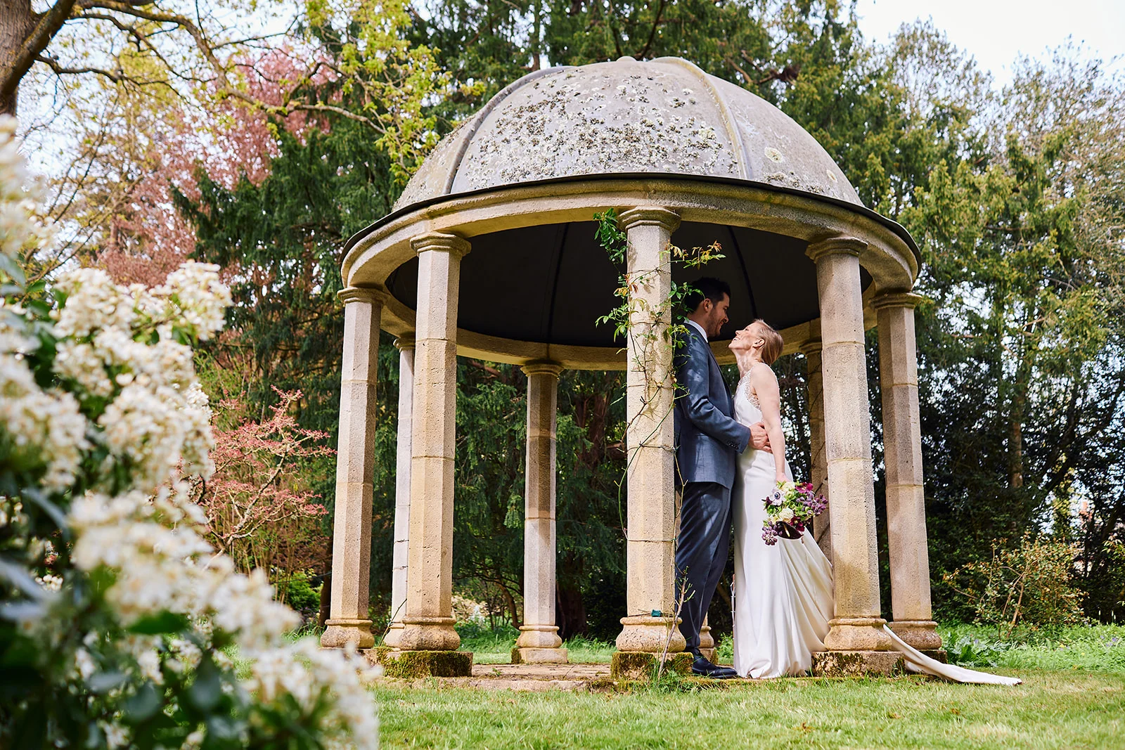 A newlywed couple stands inside a stone garden folly surrounded by lush spring greenery, holding a bouquet and looking lovingly at each other.